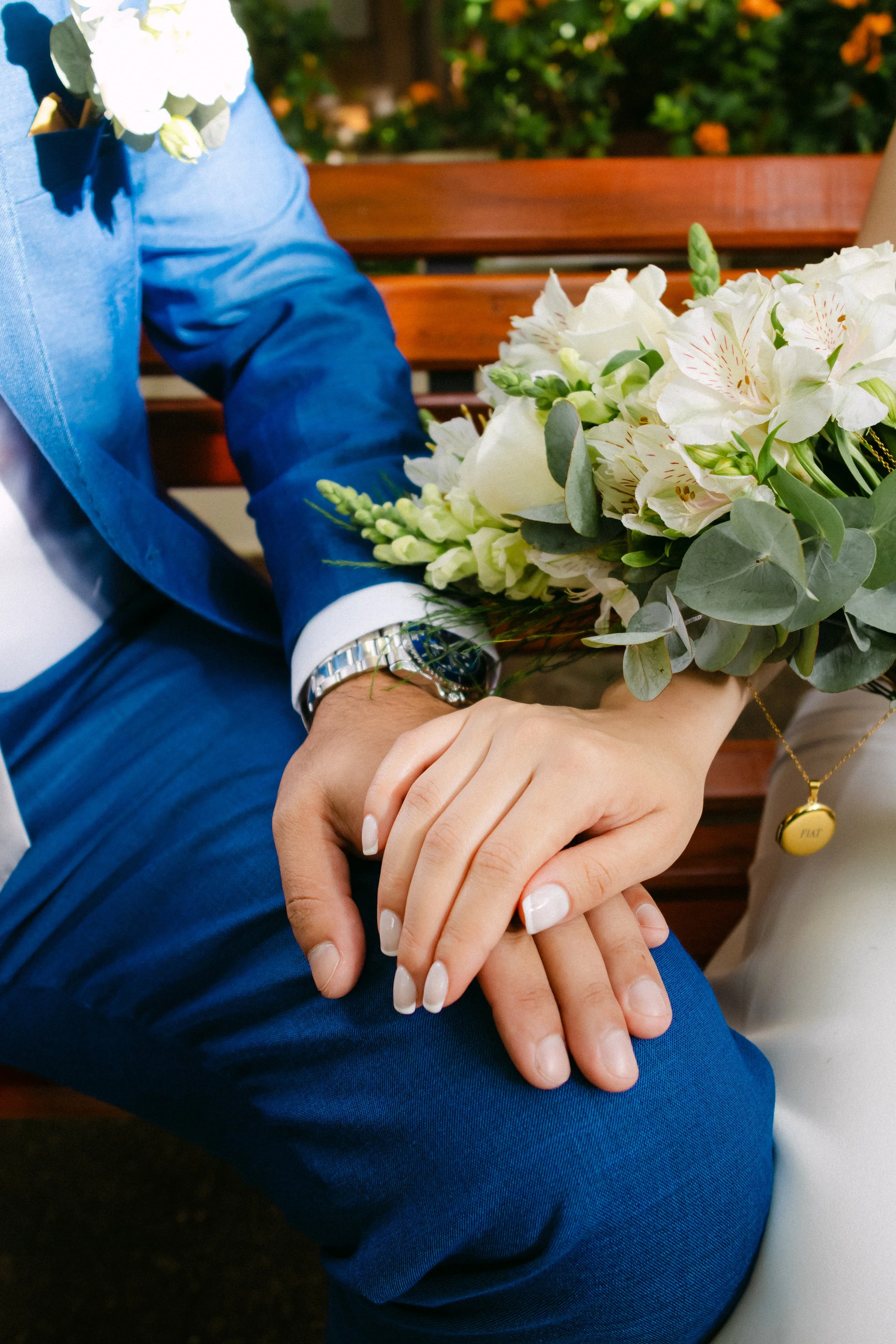 Mano de un hombre con reloj sujetando la mano de una mujer con uñas pintadas, ambos sosteniendo un ramo de flores blancas en un entorno de parque