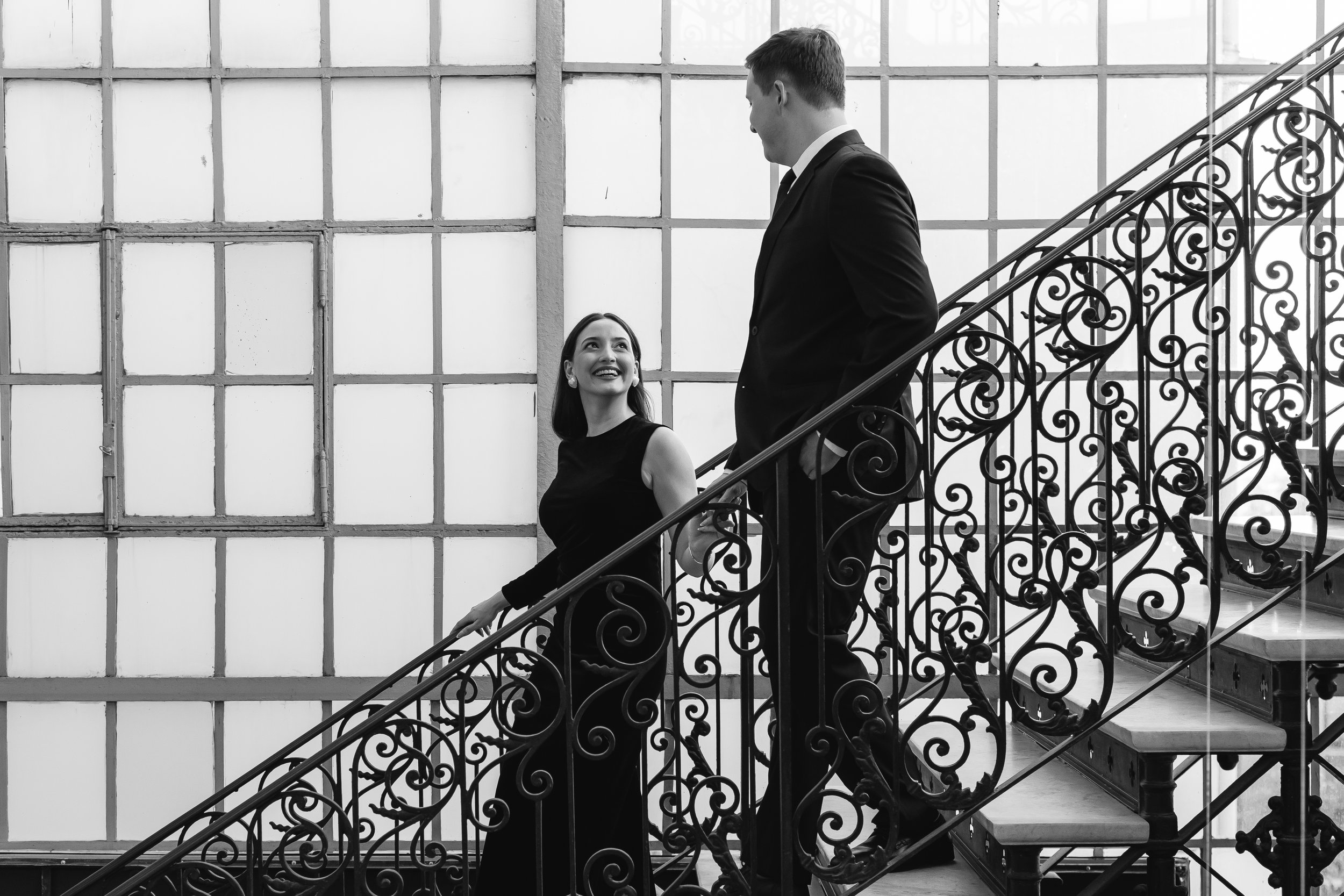 Pareja conversando en una escalera interior con barandilla de hierro decorativo, en un ambiente moderno con paredes de ventanas grandes.