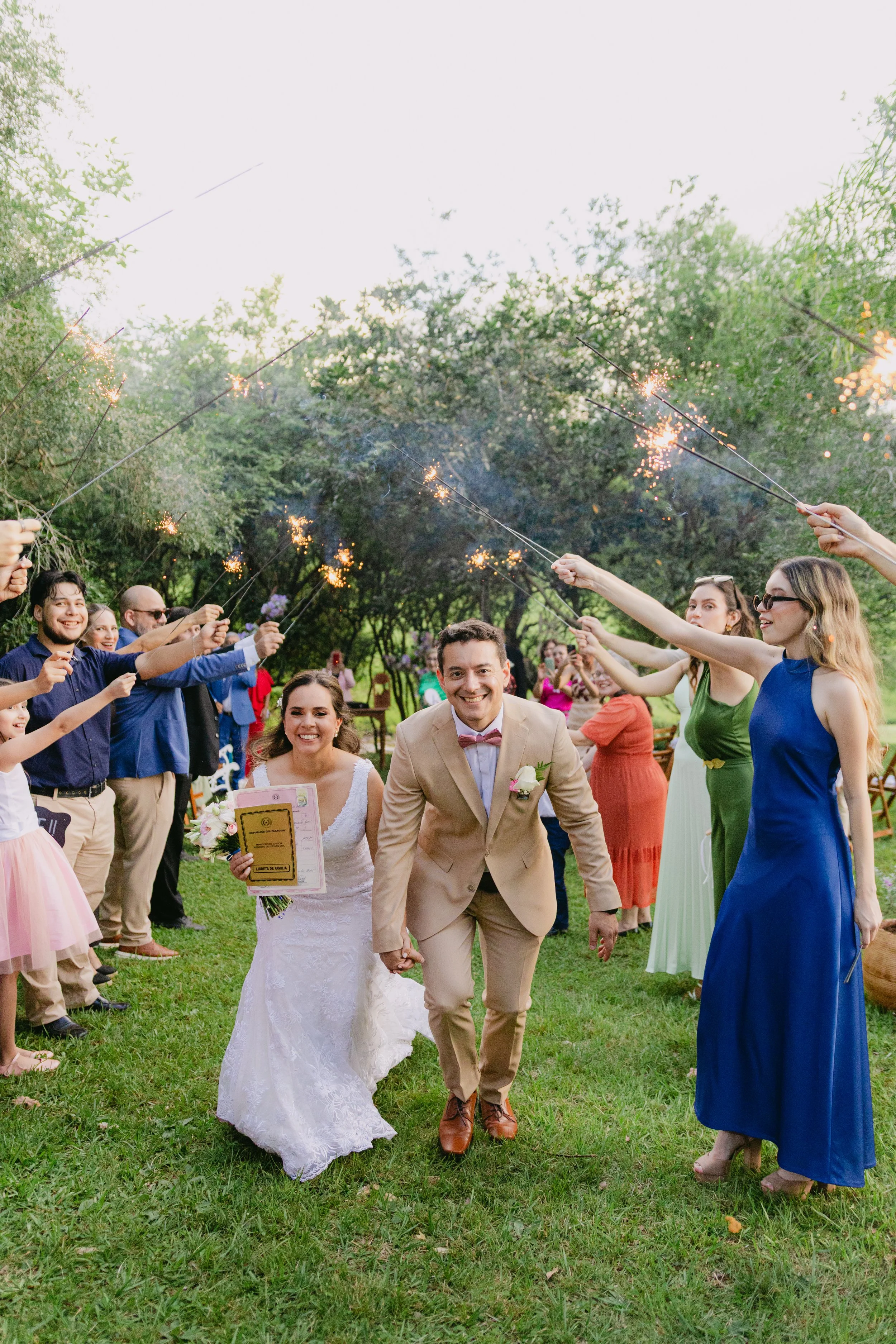 Una pareja de recién casados caminando felizmente entre amigos y familiares en una celebración al aire libre, rodeados de árboles, con personas sosteniendo bengalas encendidas, en un ambiente festivo y alegre.