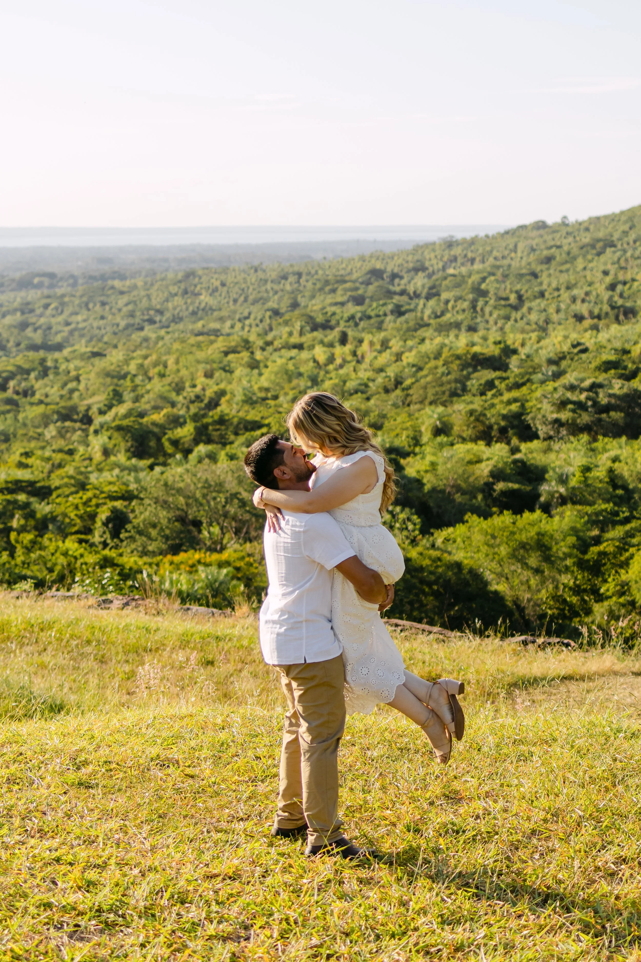 Pareja romántica en un campo verde, con un paisaje de bosques y colinas al fondo, durante el día.
