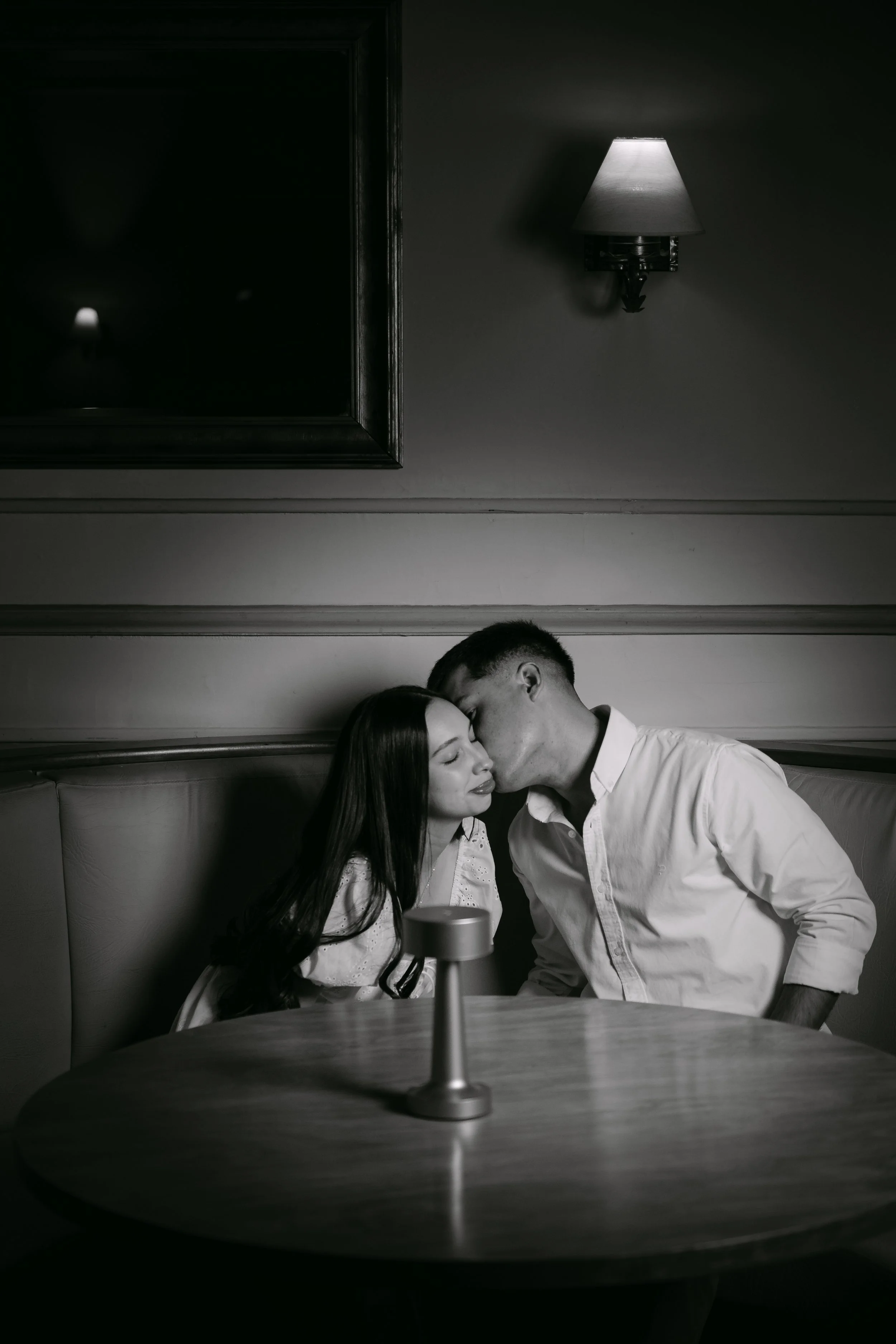 Pareja de jovenes en silencio en un restaurante, ella con ojos cerrados, el le besa en la frente, en un ambiente de luz tenue en blanco y negro.