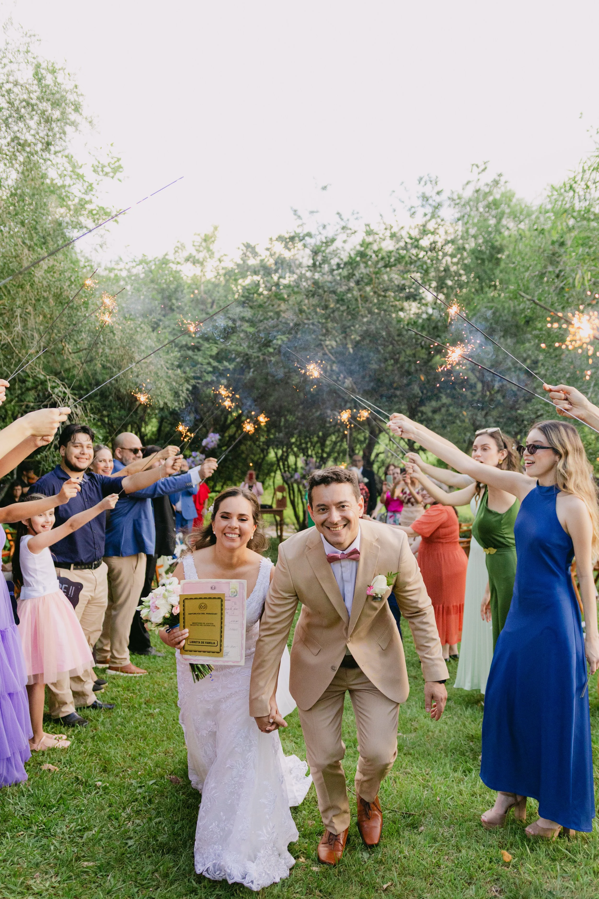 Pareja de recién casados caminando por un pasillo formado por amigos y familiares que sostienen bengalas en un entorno al aire libre con árboles y césped.