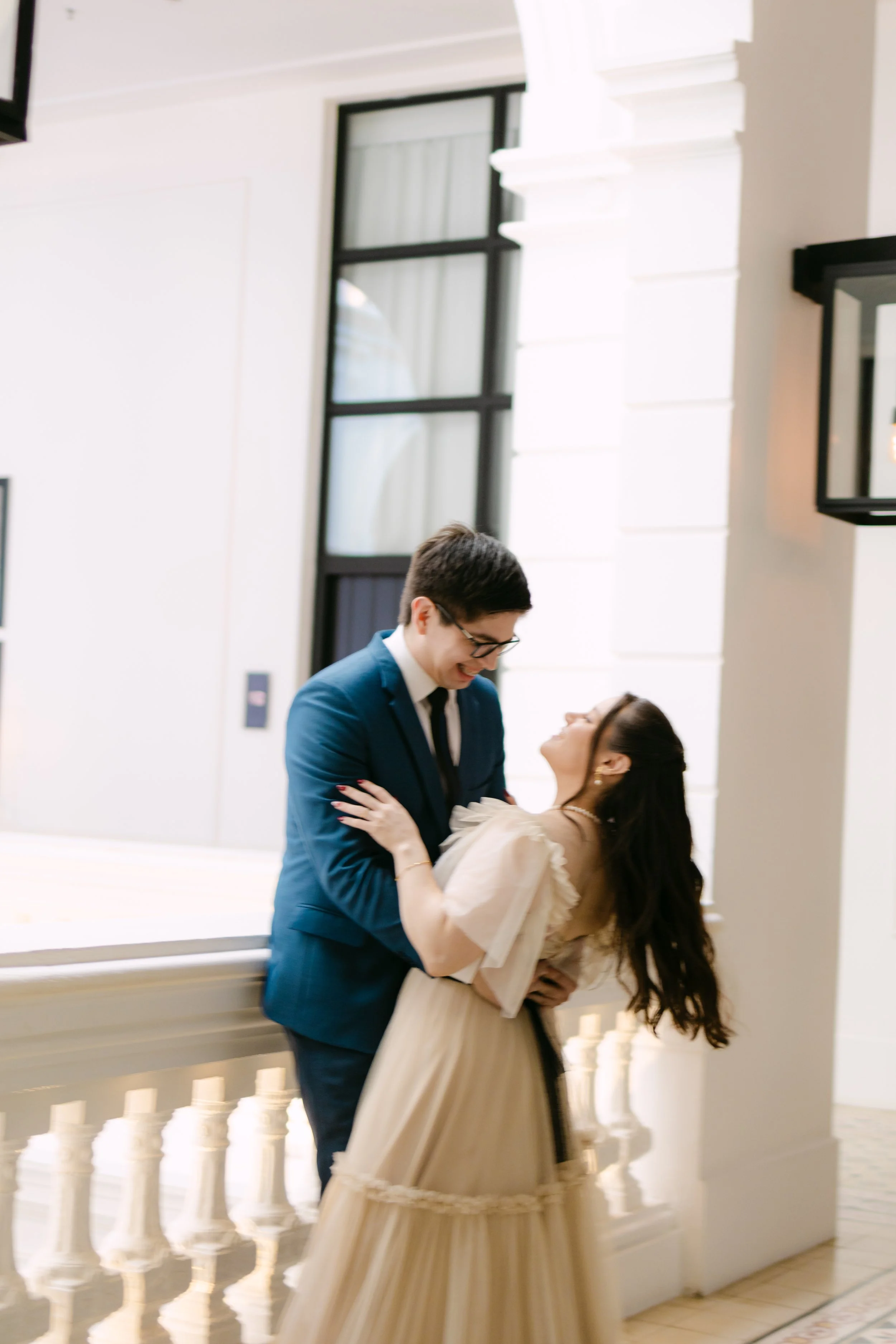 Una pareja joven en ropa elegante, sonriendo y bailando en un lugar con paredes blancas y ventanas grandes, en un ambiente que parece de celebración o boda.