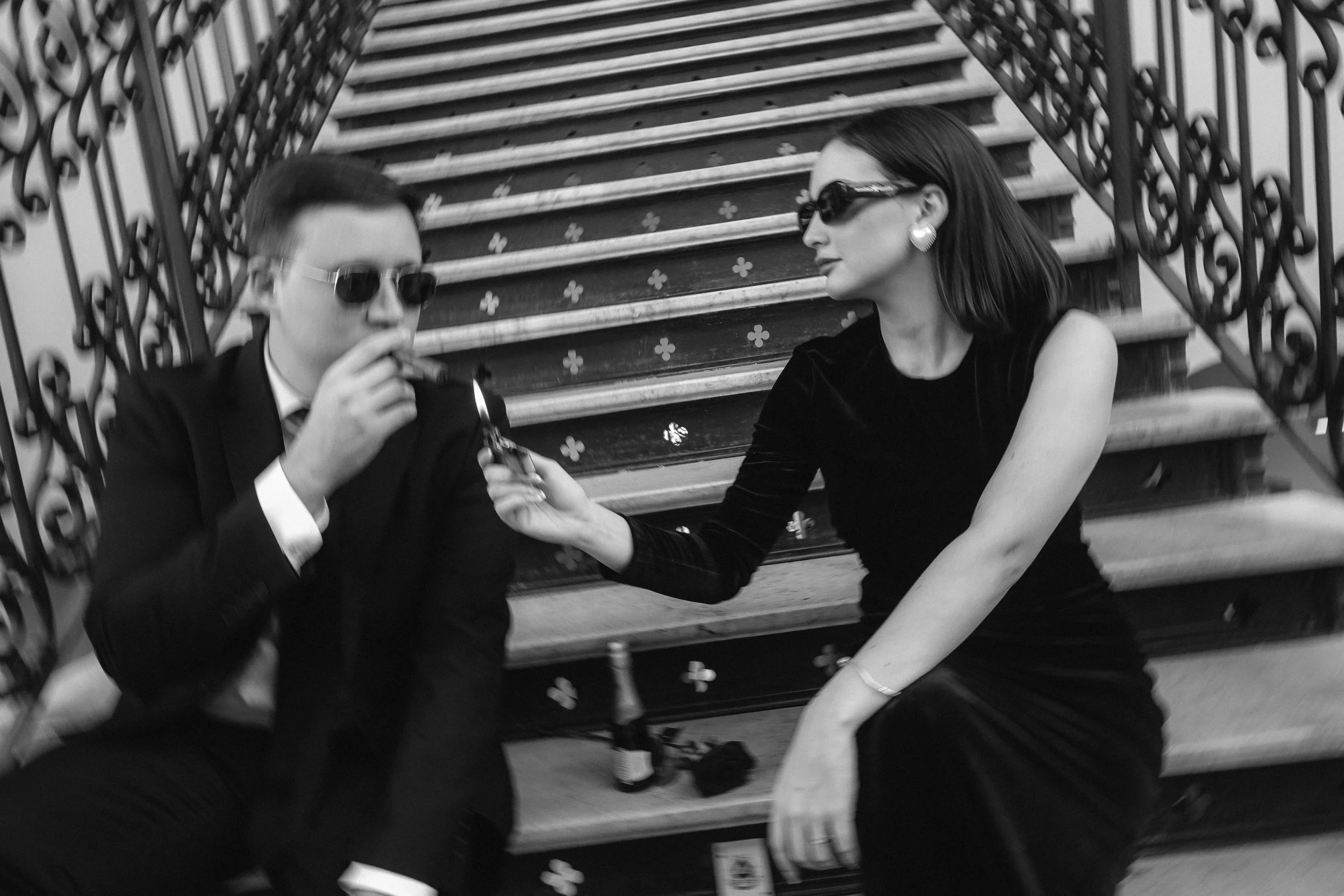 Dos personas, hombres y mujeres, con gafas de sol sentados en las escaleras, una de ellas le pasa un encendedor a la otra.