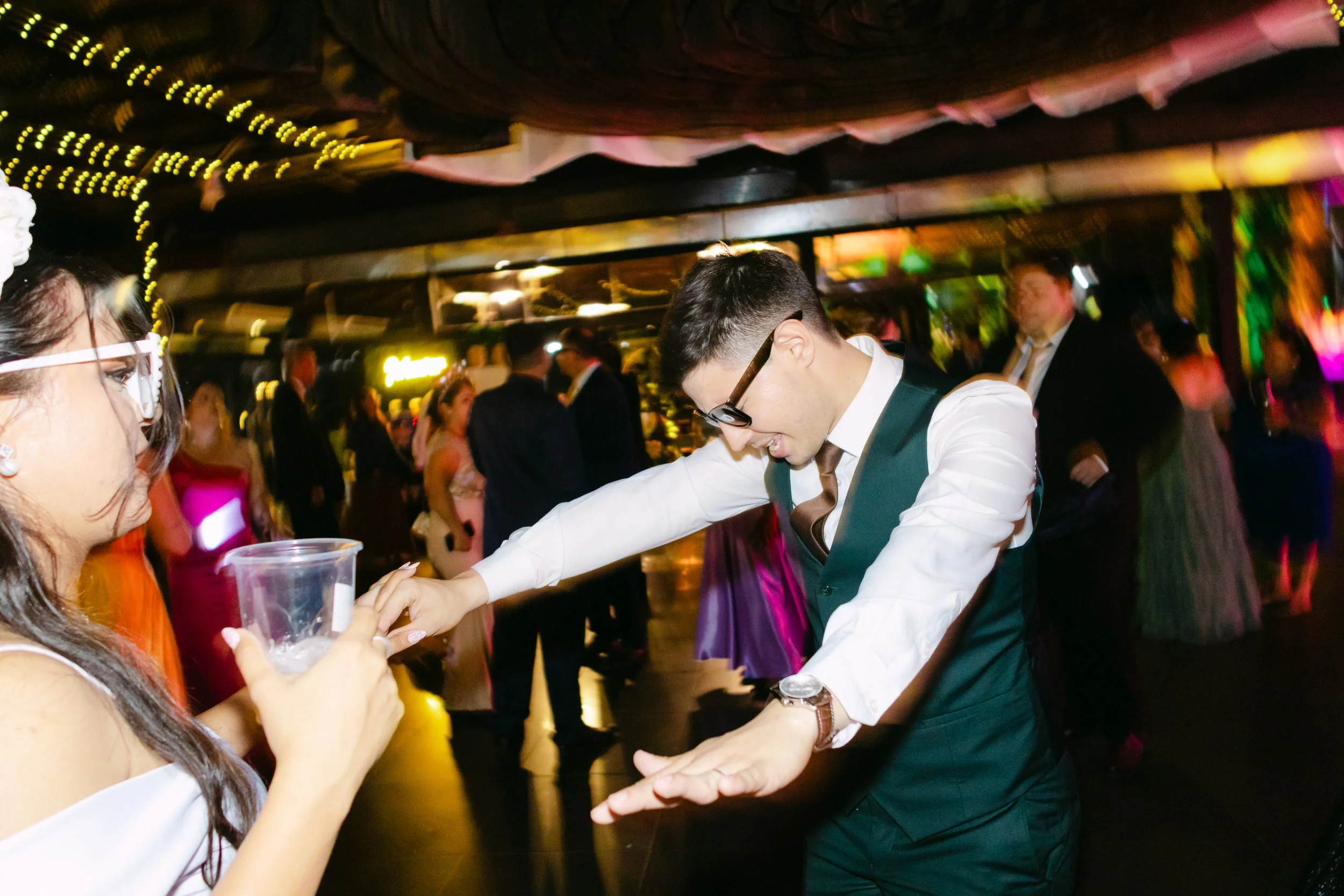 Joven bailando en una fiesta elegante con muchas personas en el fondo, luces de colores y decoración festiva.