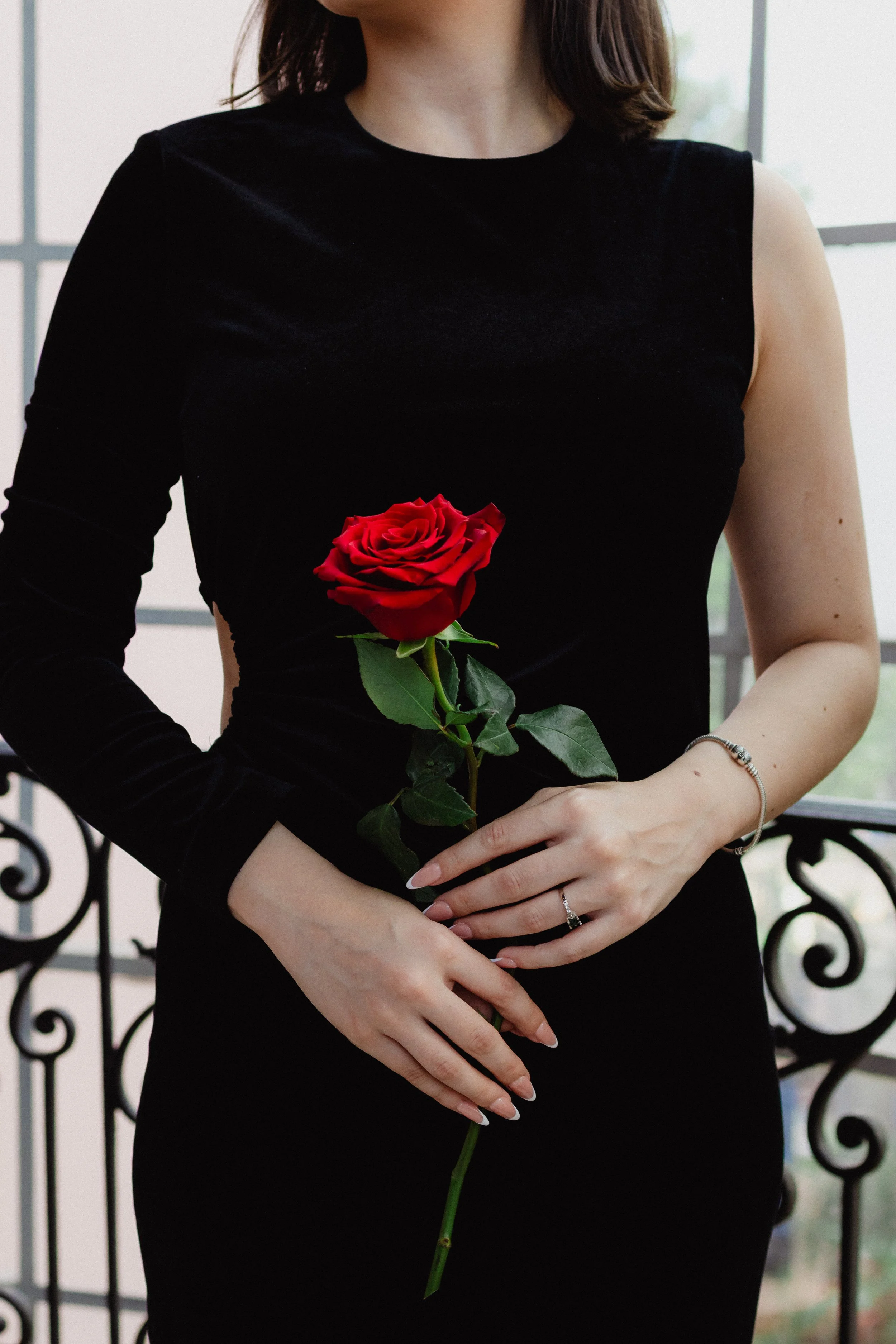Mujer con vestido negro sosteniendo una rosa roja en ambas manos, de fondo un balcón con barandilla de hierro.