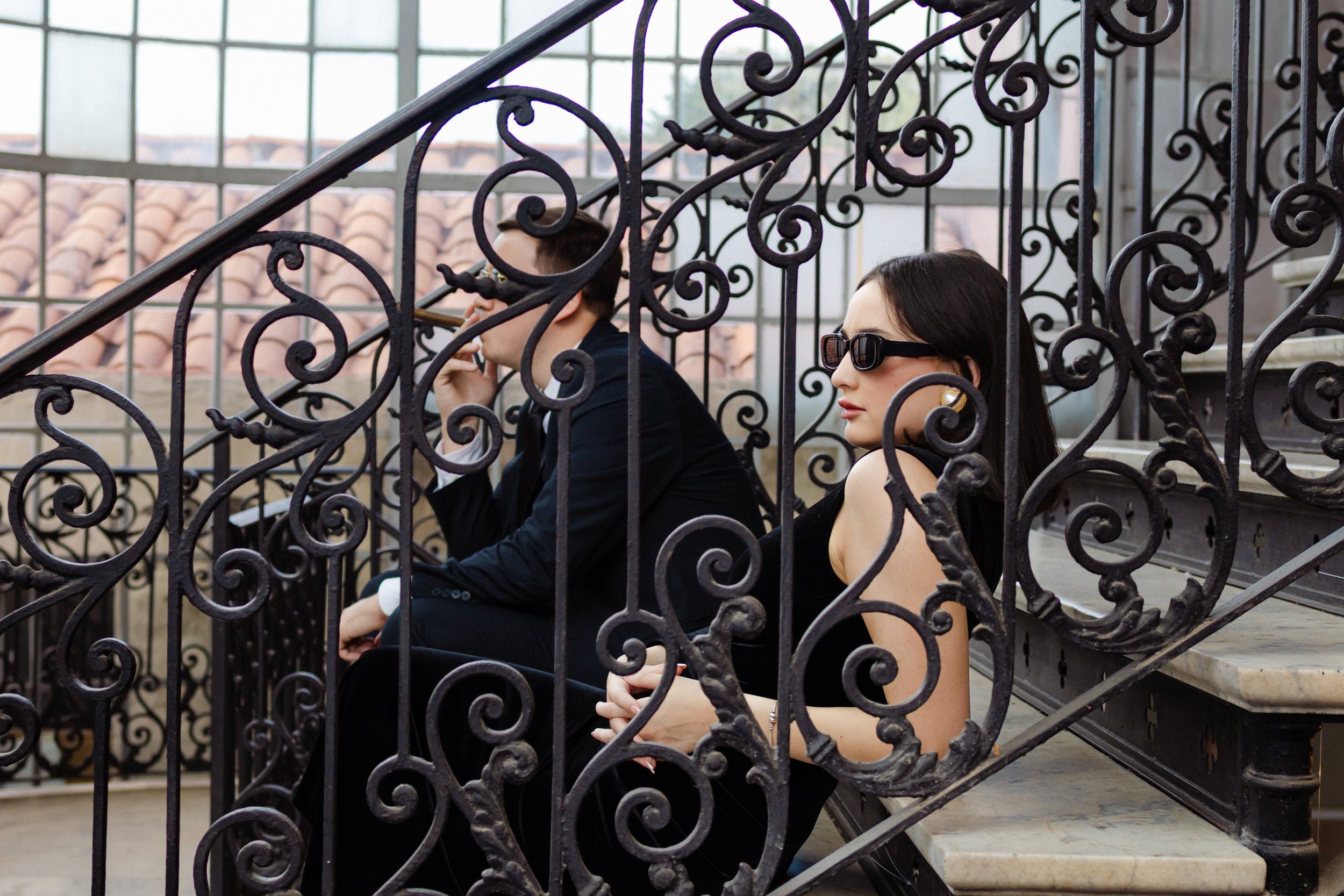 Una mujer y un hombre sentados en las escaleras de un balcón de hierro, ella usando gafas de sol y un vestido negro, él con traje oscuro y fumando.