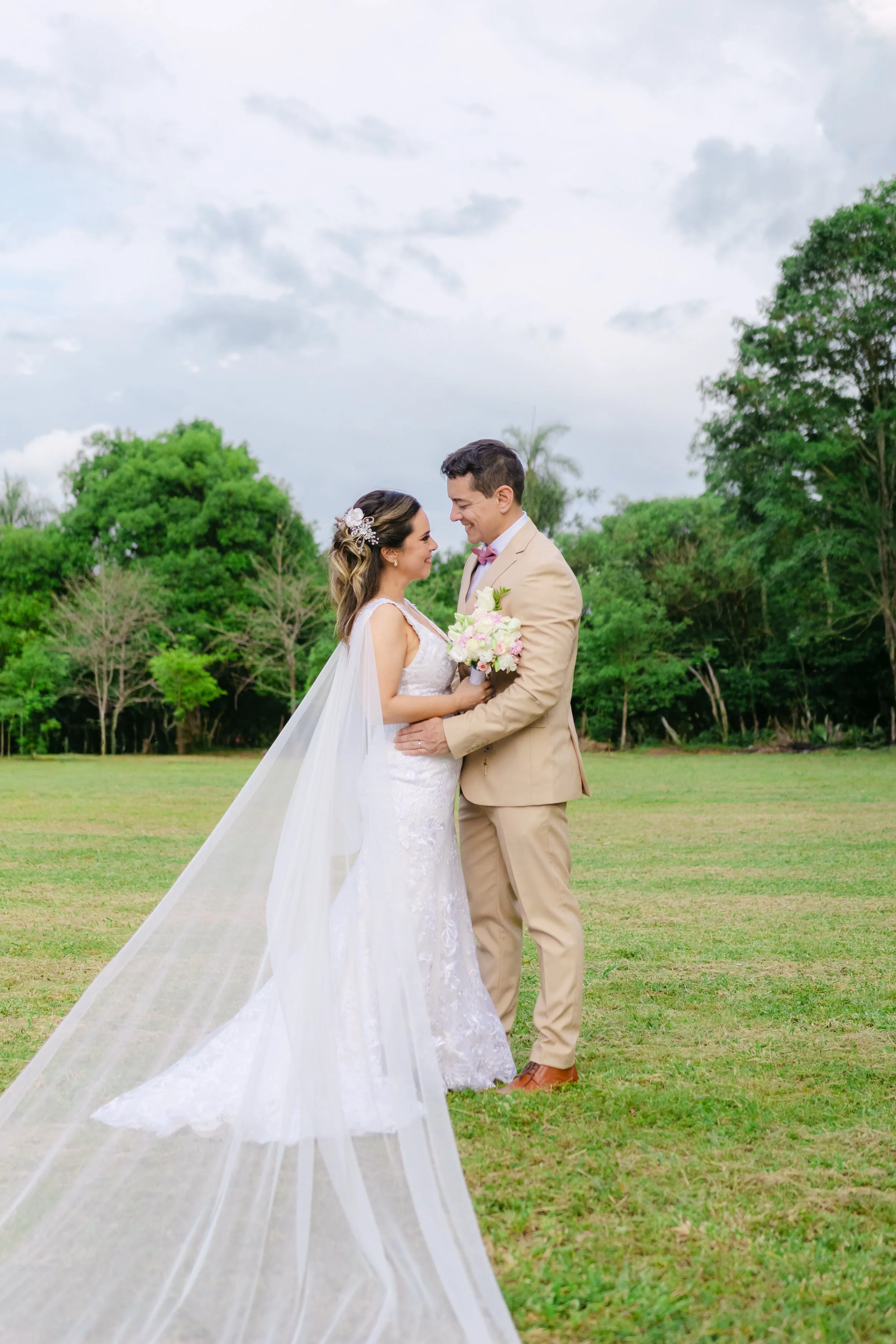 Una pareja de novios en un campo abierto, ella con vestido de novia y velo largo, él con traje beige, se miran y sonríen, rodeados de árboles y nubes en el cielo.