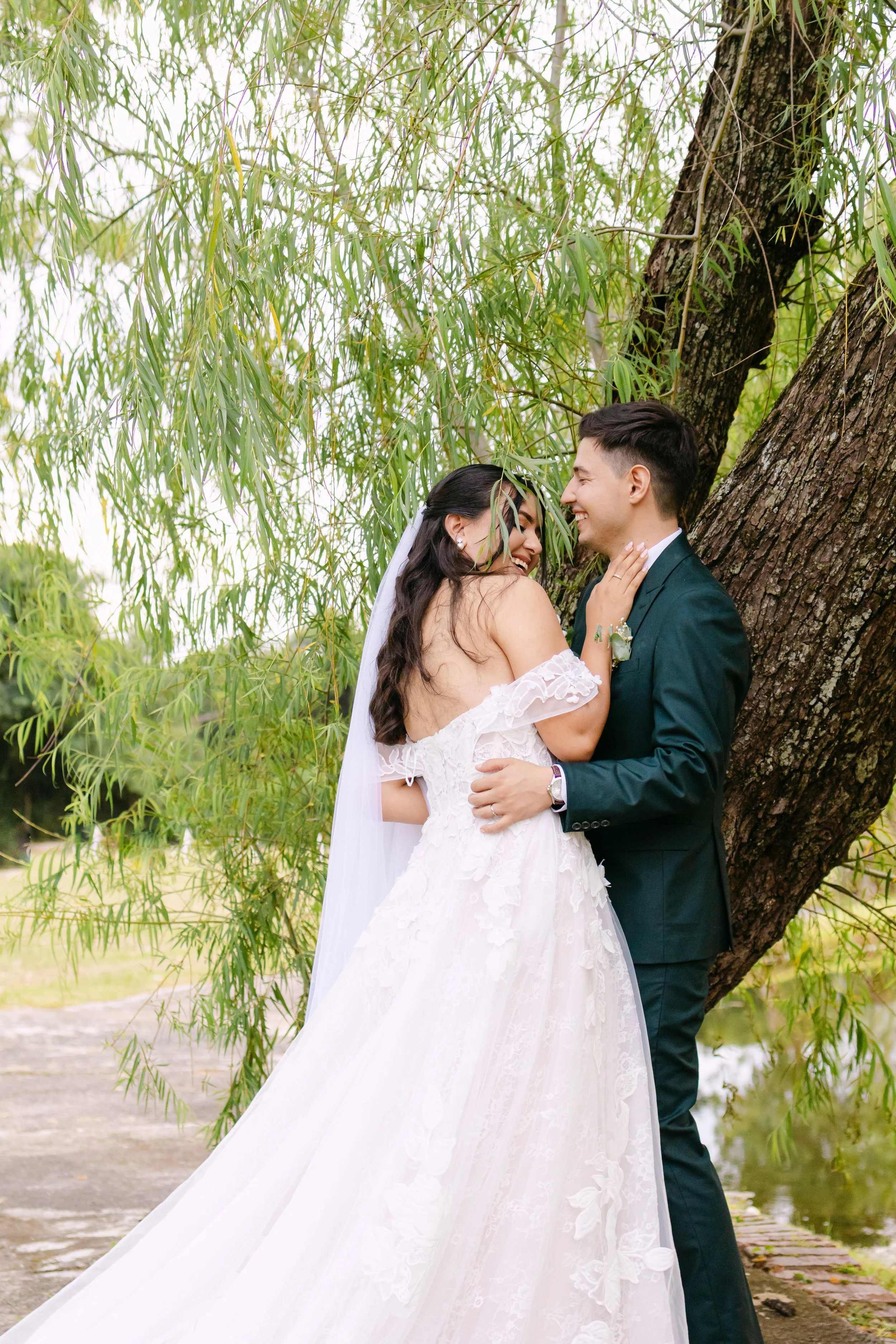 Pareja de novios en su boda, abrazados y sonriendo, al aire libre junto a un árbol grande y frondoso.