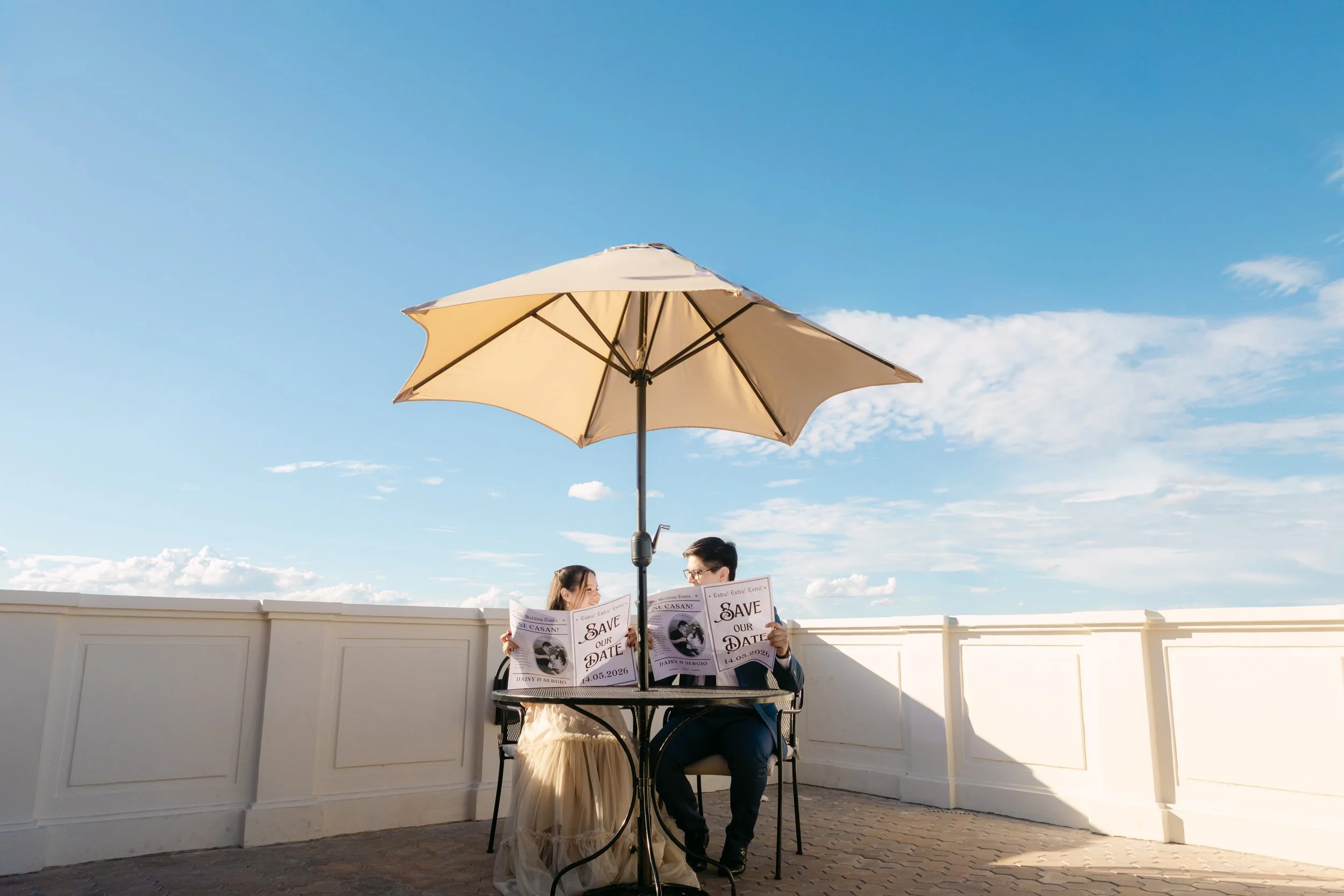 Una pareja sentada en una mesa con un paraguas en un espacio al aire libre, leyendo carteles que dicen 'Save Our Date 14.05.2026'.