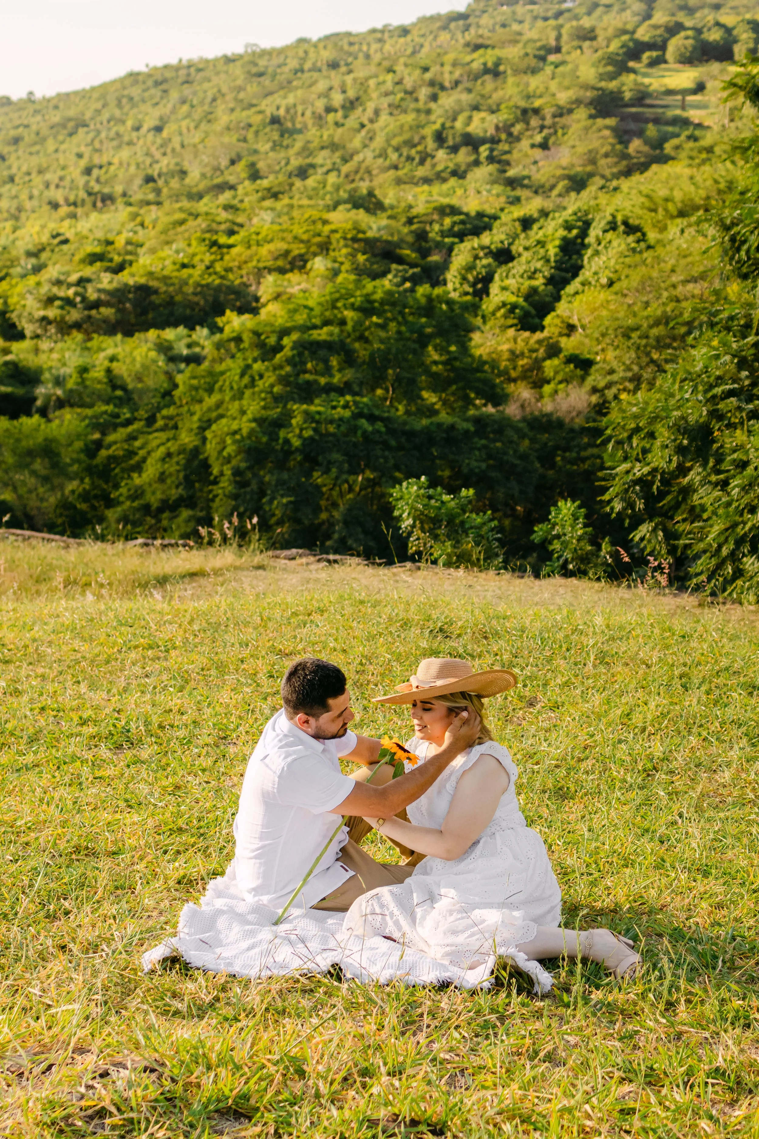 Una pareja en ropa blanca sentada sobre el césped en un campo, rodeados de naturaleza. La mujer lleva un sombrero de paja, y el hombre le da un masaje en la cara en un día soleado.