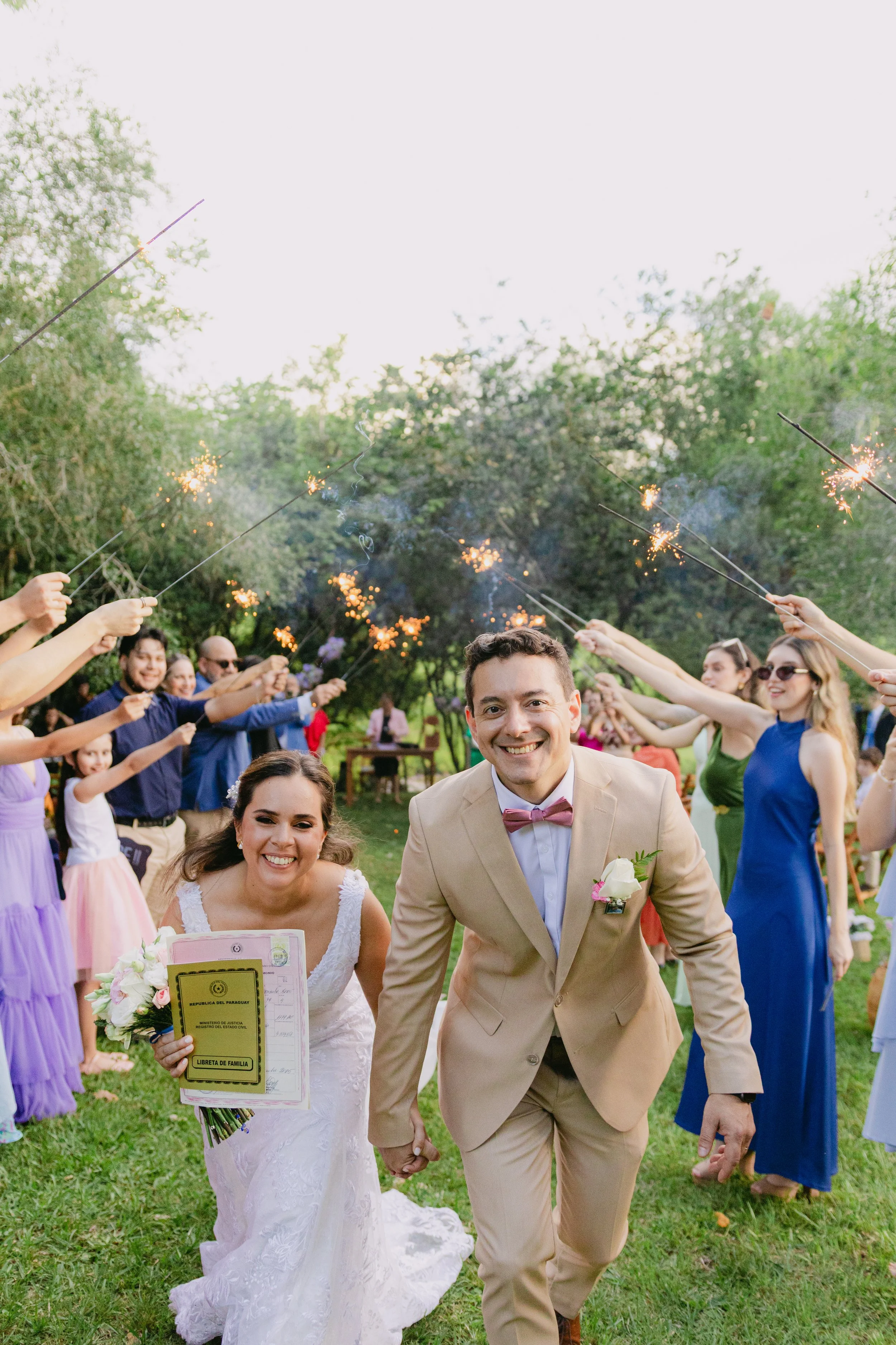 Pareja de novios en su boda, sonriendo y caminando por el césped, rodeados de invitados que lanzan bengalas y celebran en un jardín al aire libre.