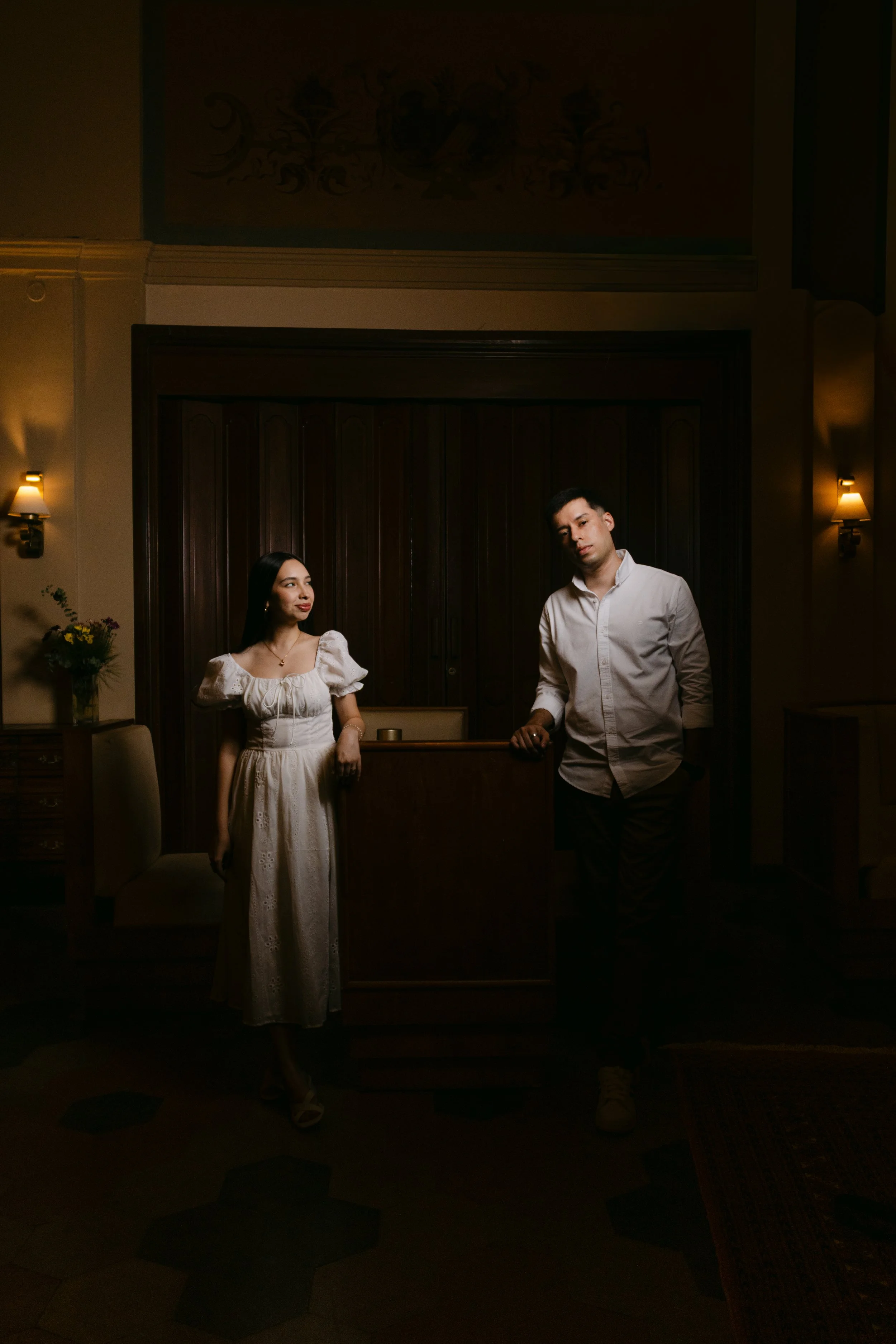 Una pareja joven en un ambiente elegante con paredes de madera oscura y lámparas de pared, una mujer con vestido blanco y un hombre con camisa blanca, en una habitación con decoración clásica y flores en un jarrón.