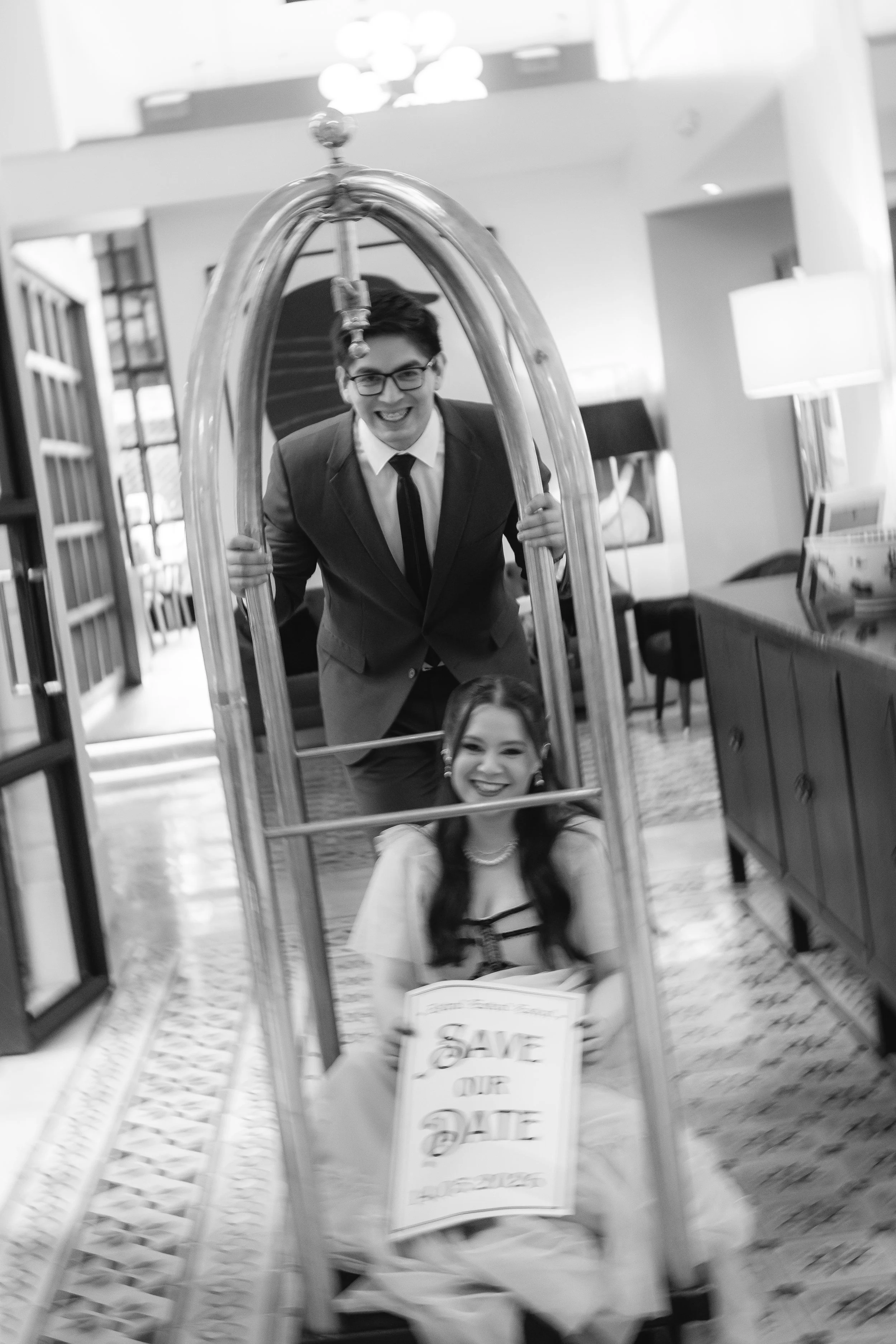 Una pareja de jovenes en un sillon con una estructura metálica, la mujer sostiene un cartel que dice 'Save Our Date' y ambos parecen estar felices.