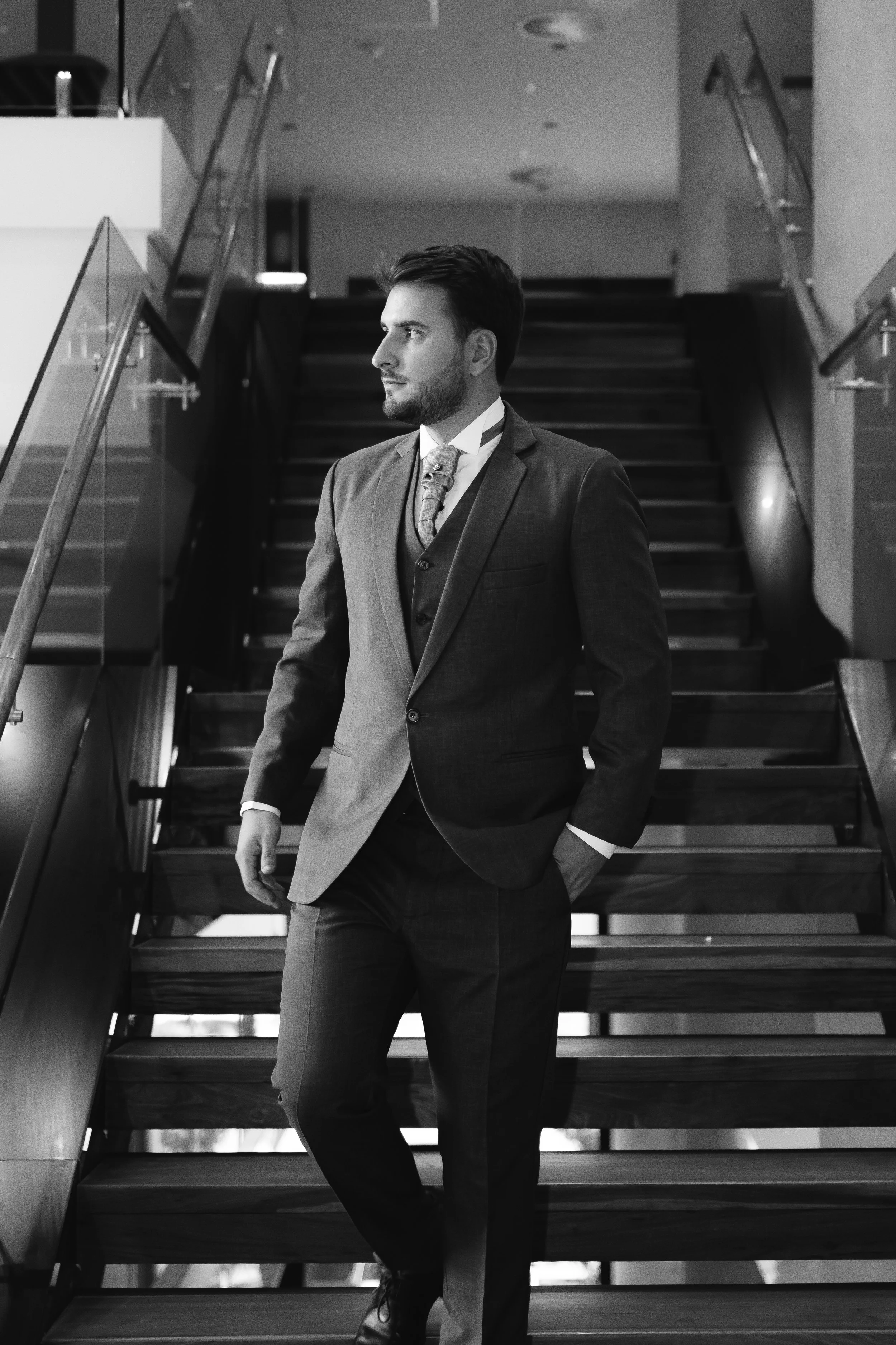 Hombre con barba y peinado peinado, vestido con saco, camisa y corbata, caminando por una escalera en un entorno moderno y elegante.