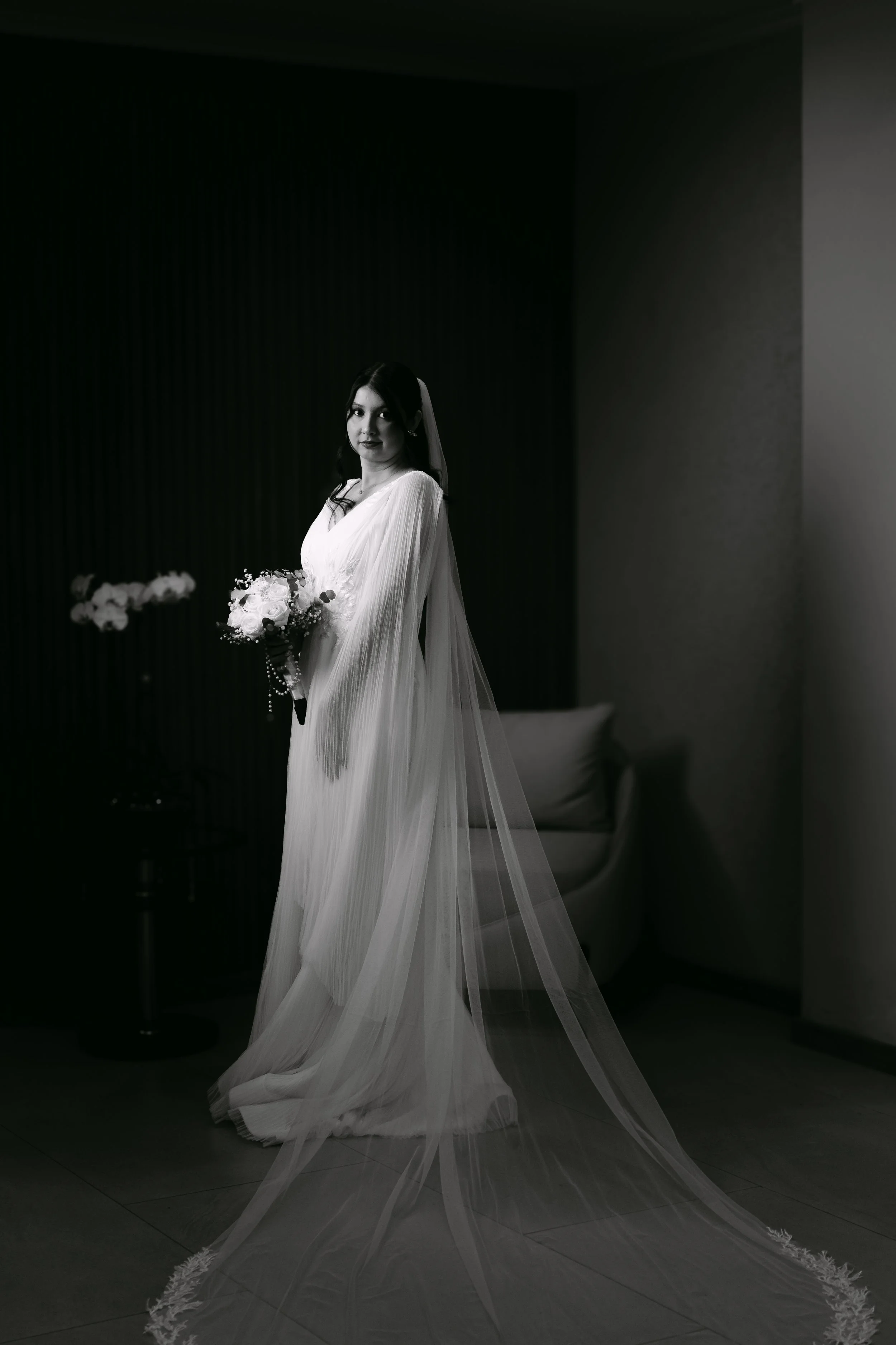 Una mujer con vestido de novia y velo, sosteniendo un ramo de flores, en un ambiente interior en escala de grises.