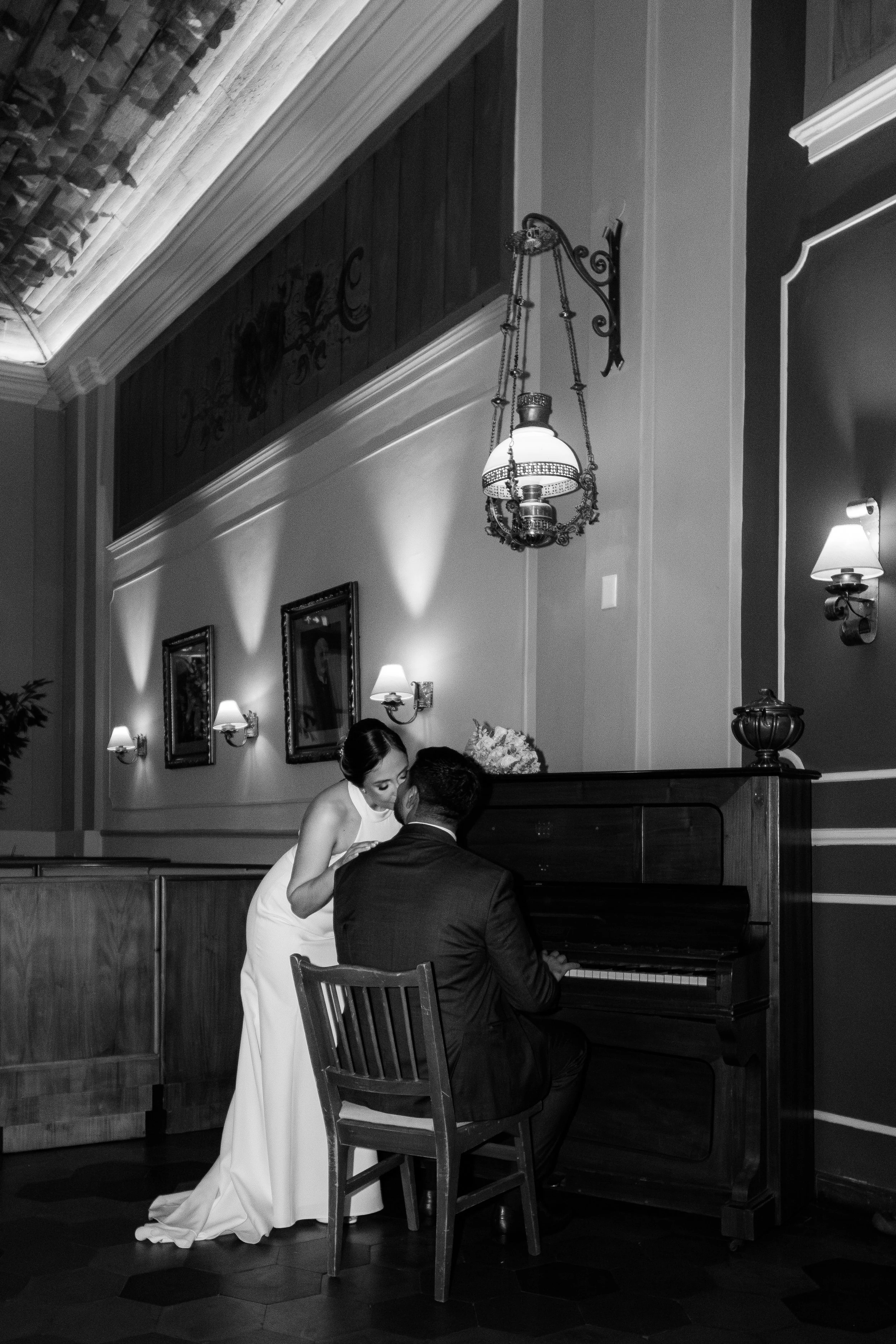 Pareja en un salón elegante, la mujer con vestido blanco y el hombre con traje oscuro, escuchando música en un piano antiguo, decorado con cuadros y lámparas en las paredes.