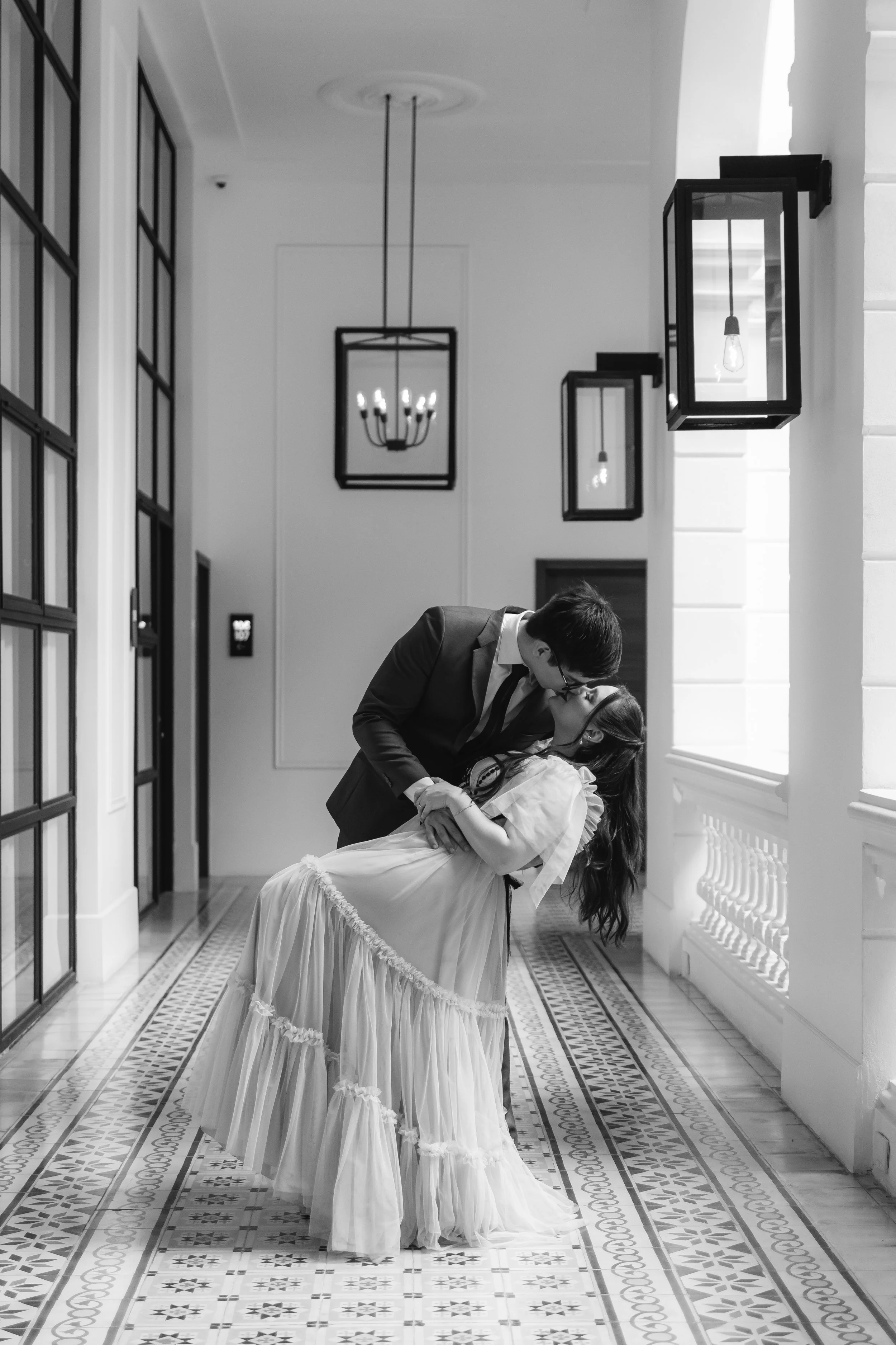 Una pareja en un pasillo, la mujer lleva un vestido largo y el hombre viste un traje, están en un momento romántico con un beso.