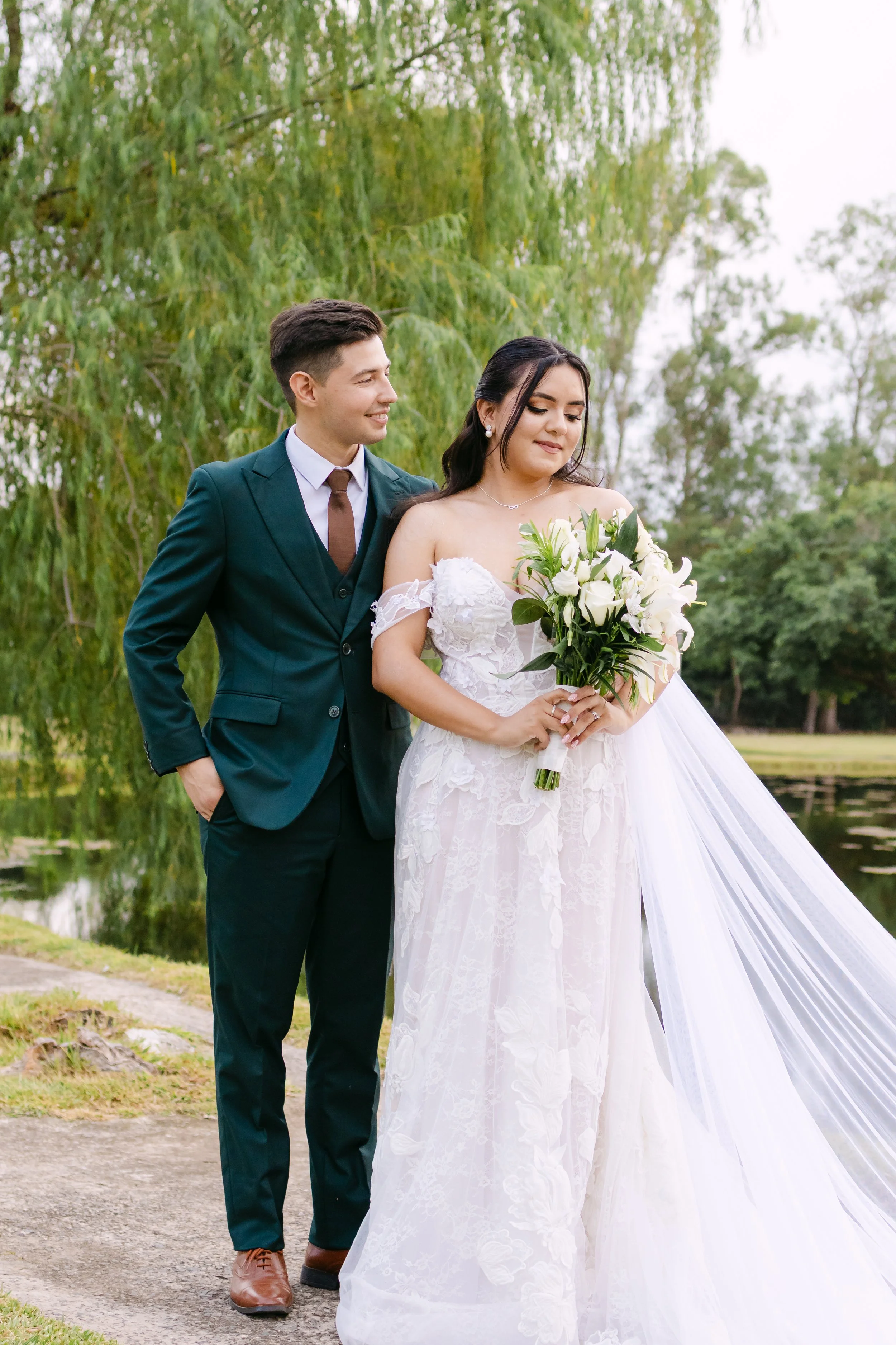 Pareja de recién casados en un parque junto a un lago, la novia con vestido de novia y ramo de flores blancas, el novio con traje oscuro y camisa blanca.