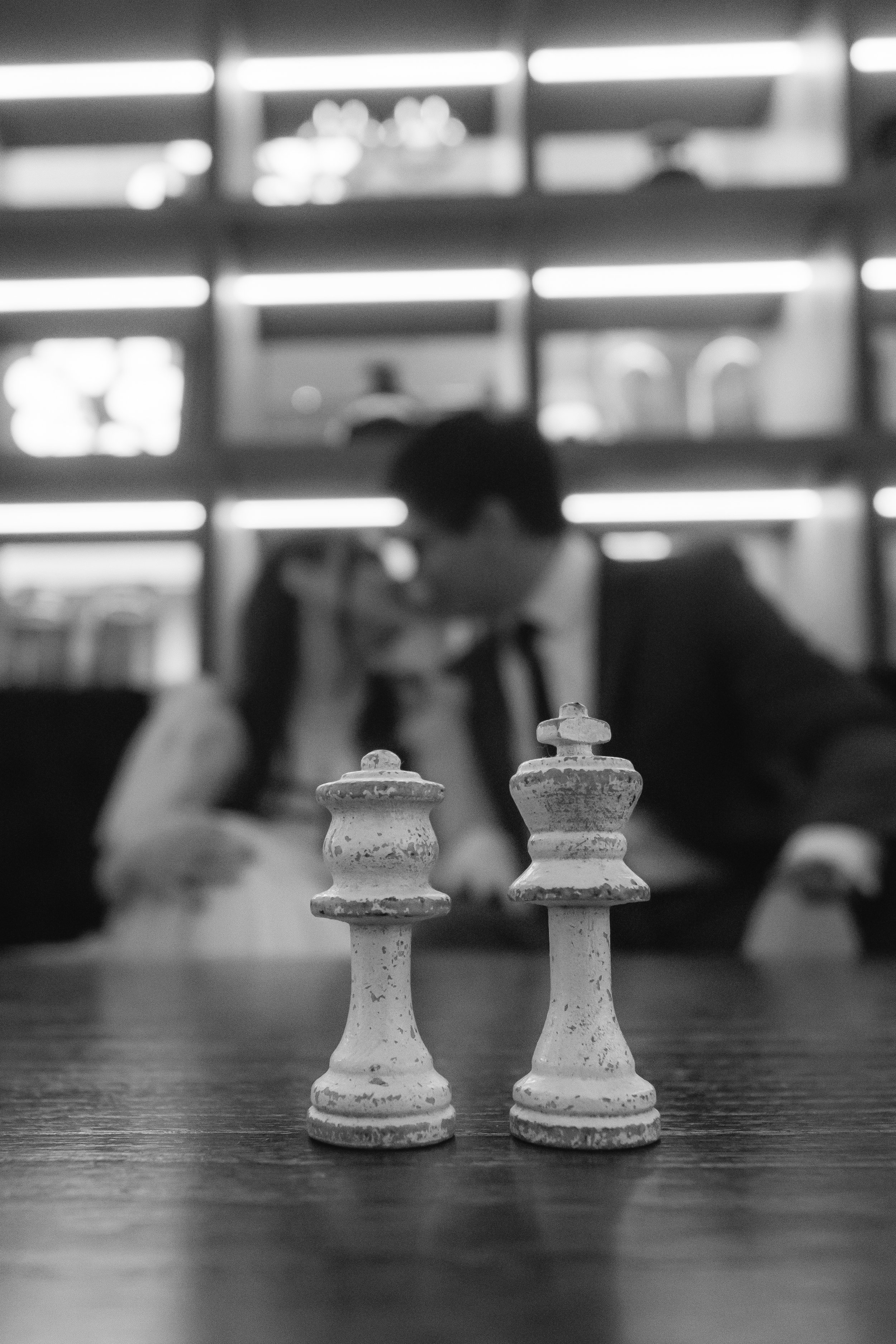 Dos piezas de ajedrez blancas en primer plano sobre una mesa, con una pareja en segundo plano en un ambiente con estanterías iluminadas.