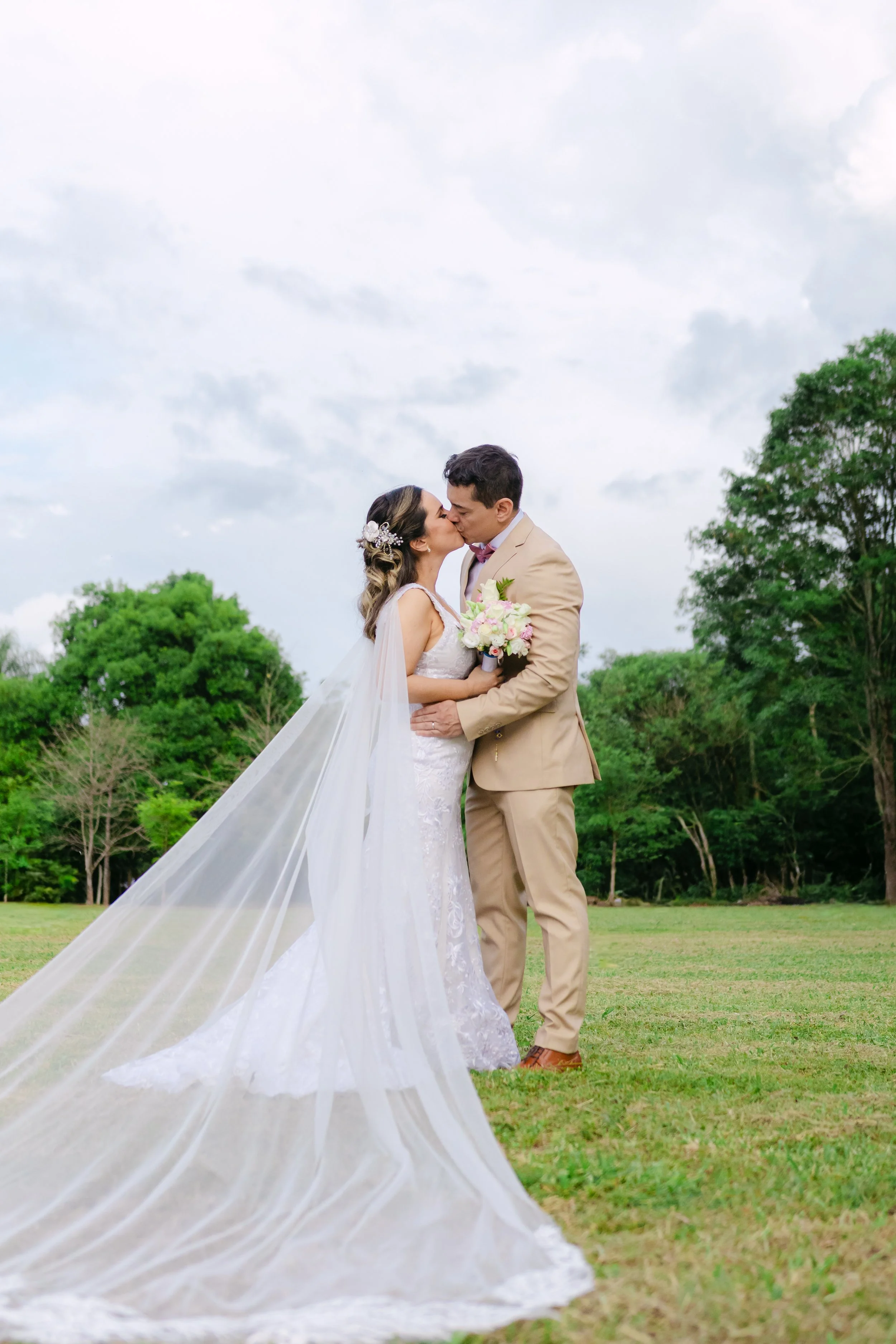 Pareja de recién casados besándose en un campo con árboles y cielo nublado, ella con vestido y velo de novia y él con traje beige, ella sostiene ramo de flores.