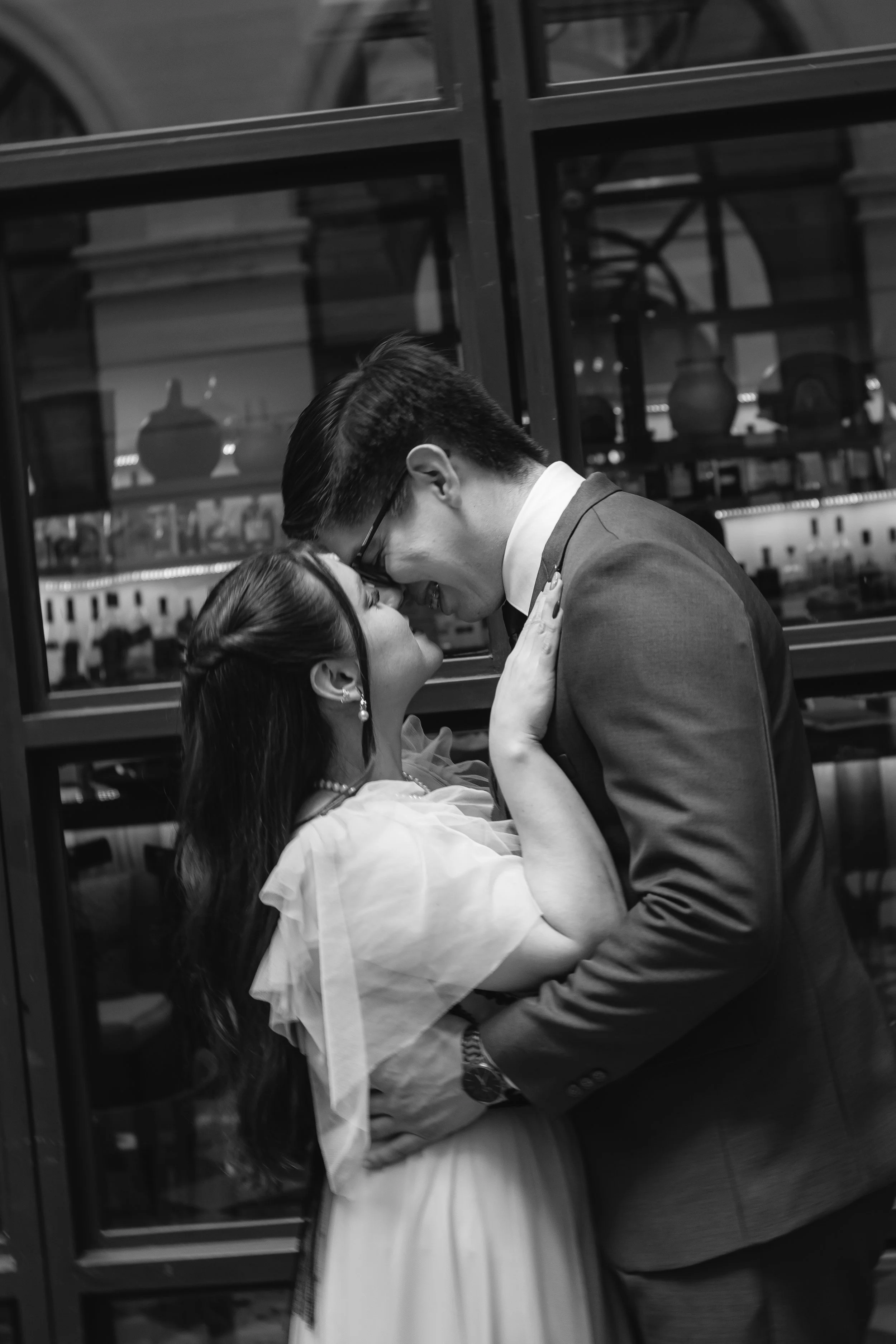 Una pareja de enamorados en un momento romántico, con la mujer sonriendo y el hombre inclinado, tocándose la frente, en un ambiente oscuro con ventanas de fondo que reflejan luces y objetos decorativos.