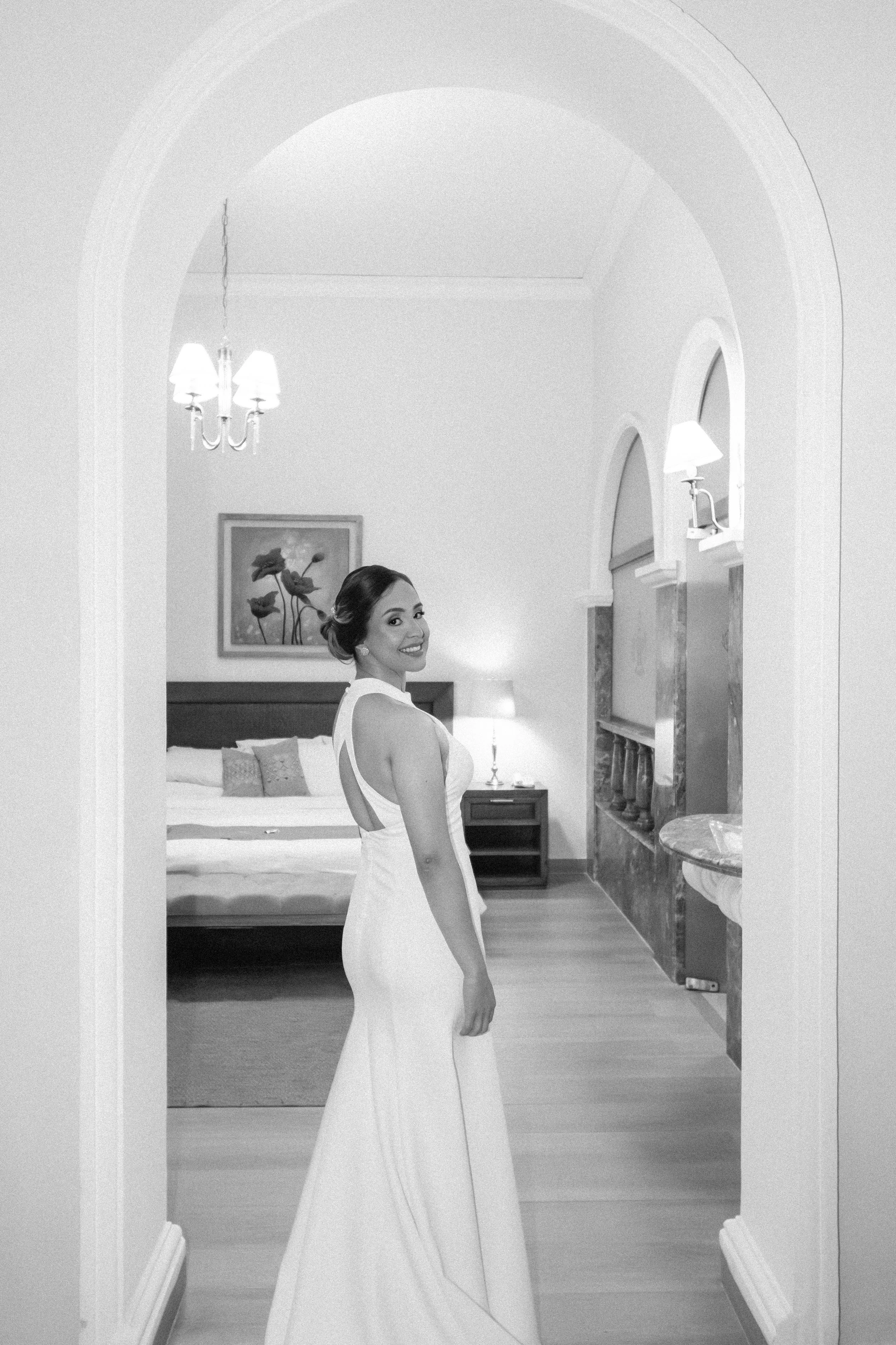Mujer sonriendo con vestido de novia en una habitación elegante, vista desde un arco en la entrada.
