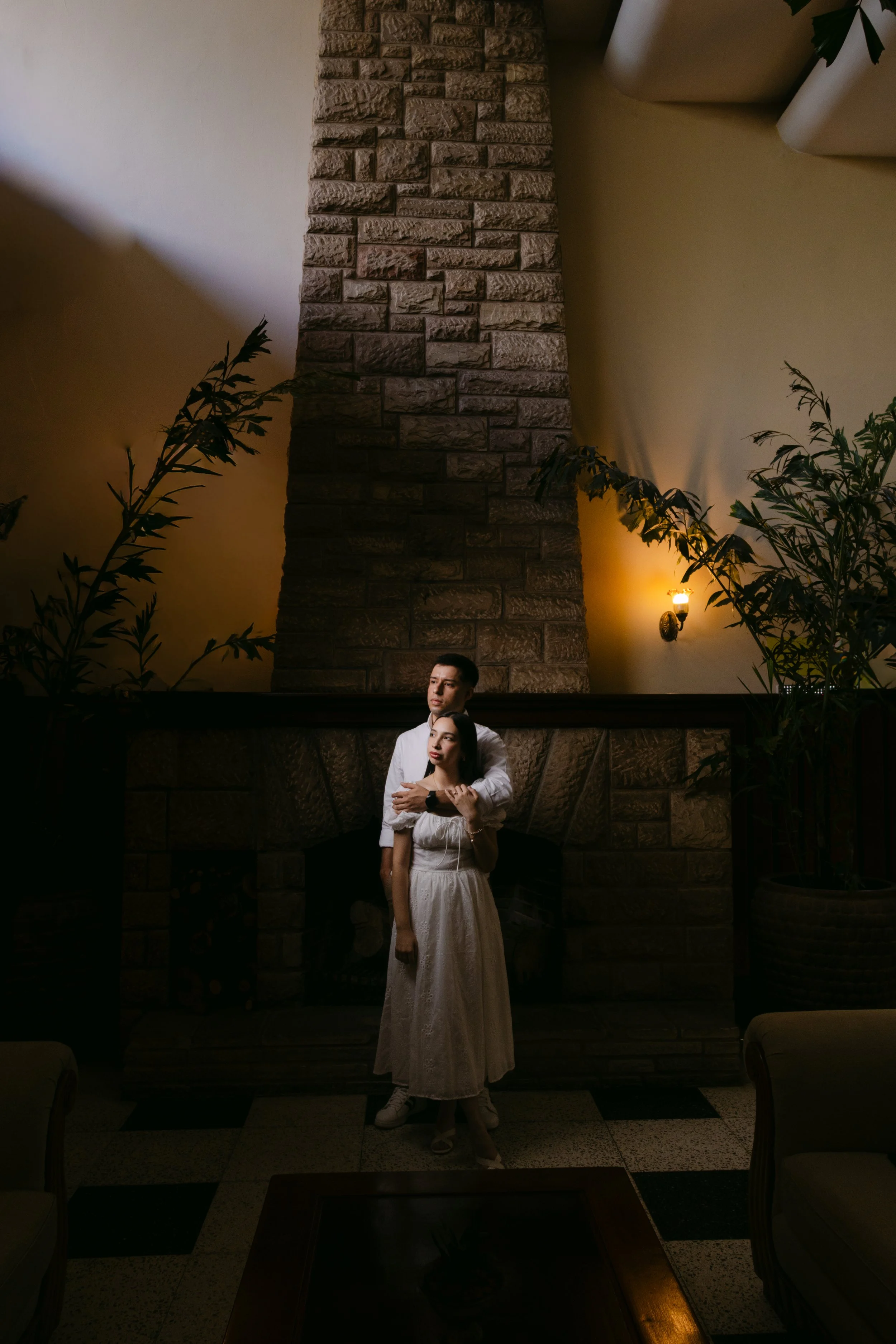 Una pareja posando en un interior con una chimenea de piedra en el fondo y una pared de color beige. La iluminación es suave y cálida.