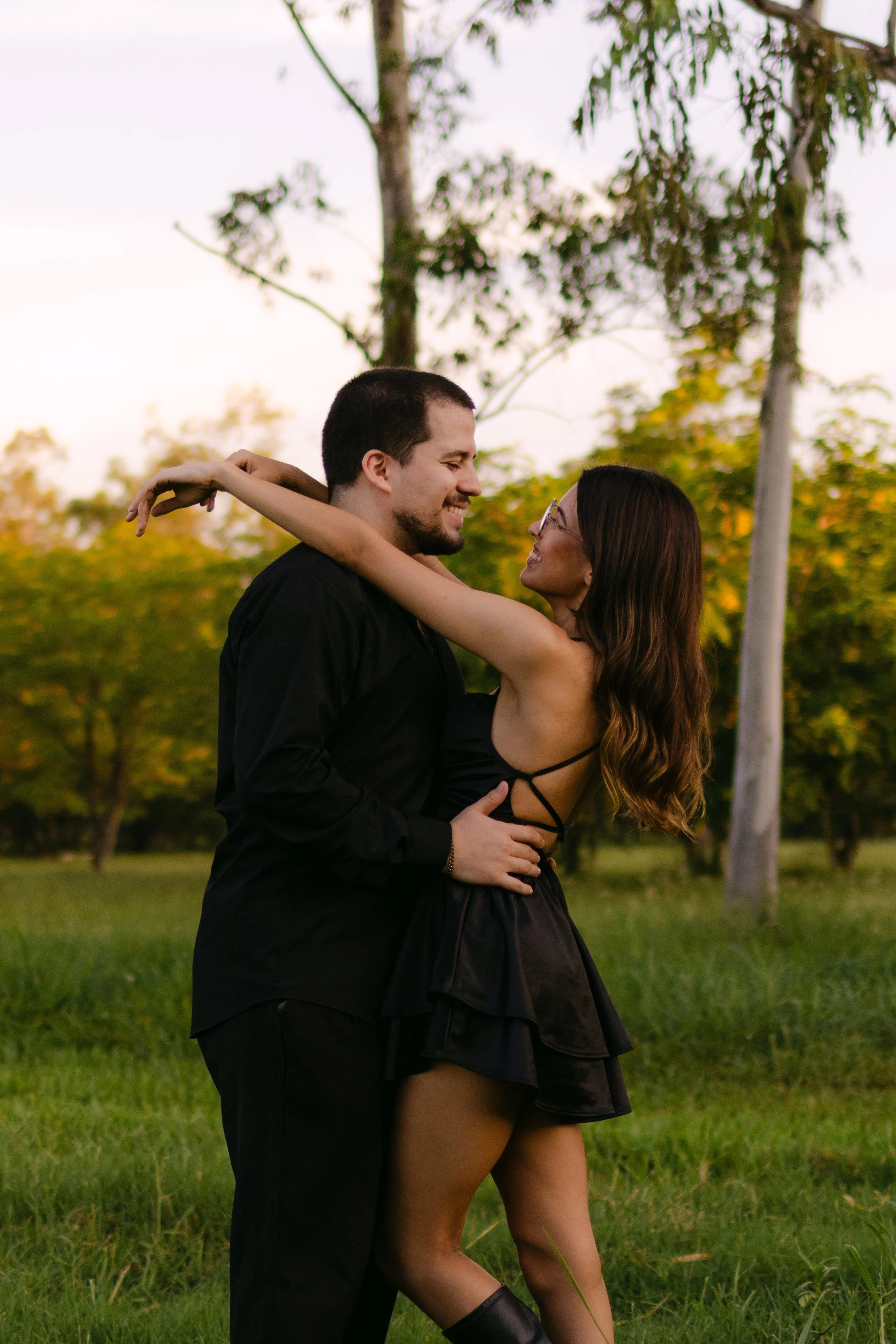 Una pareja bailando en un parque, rodeados de árboles y césped, durante el atardecer, con expresiones de felicidad.