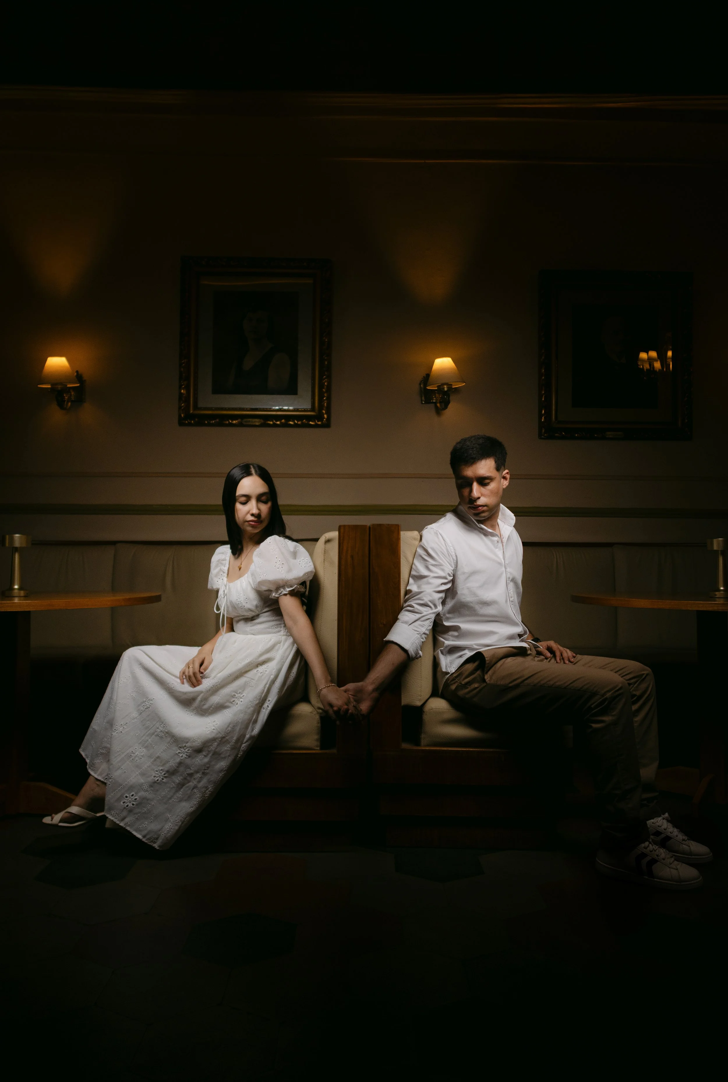 Una pareja está sentada en un banco, la mujer con vestido blanco y el hombre con camisa blanca y pantalones beige, sosteniéndose de las manos y mirando en direcciones opuestas, en un ambiente de interior con paredes decoradas y cuadros.