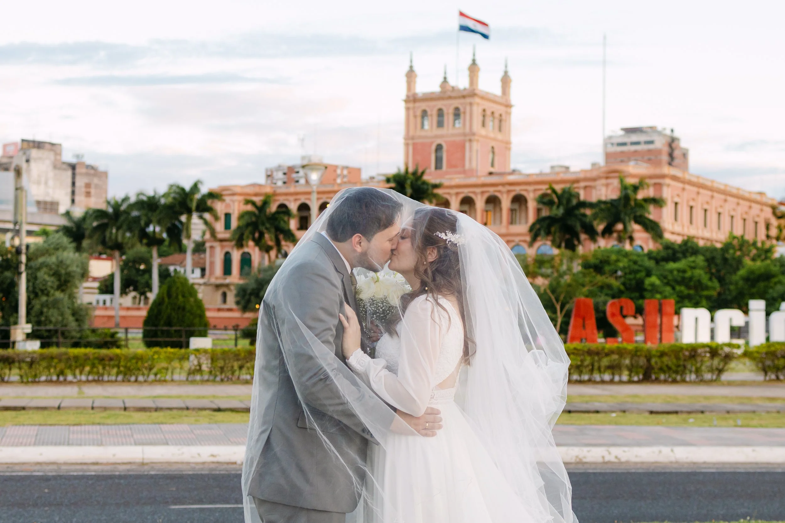 Una pareja casada besándose con un velo de novia, fondo de edificio rosado y banderas, en la ciudad de Asunción, Paraguay.