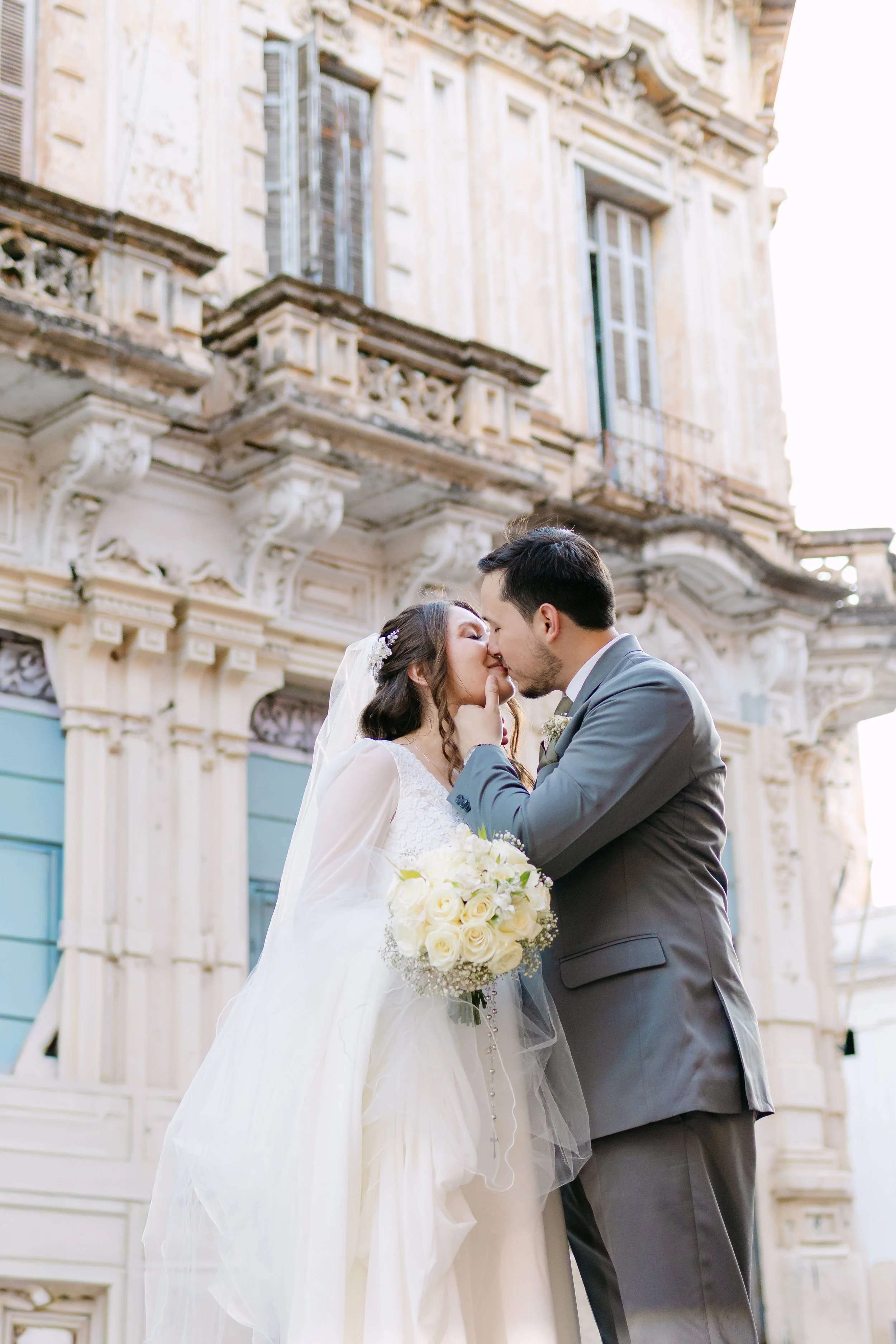 Una pareja de recién casados se besa frente a un edificio antiguo con detalles ornamentales.