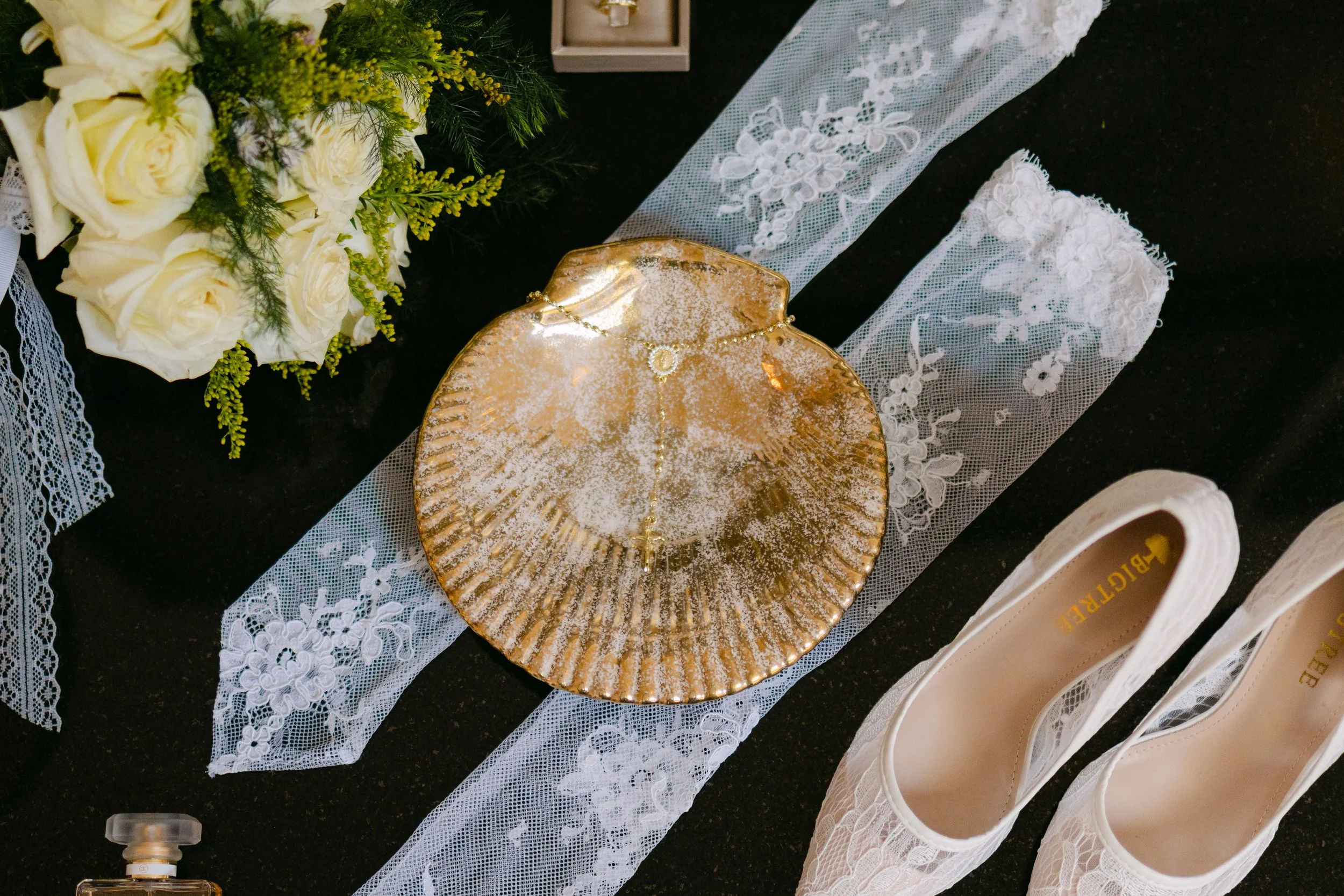Flores blancas decorativas, una cadena de oro en un plato dorado, y zapatos beige sobre una tela de encaje blanco, todo sobre una superficie negra.