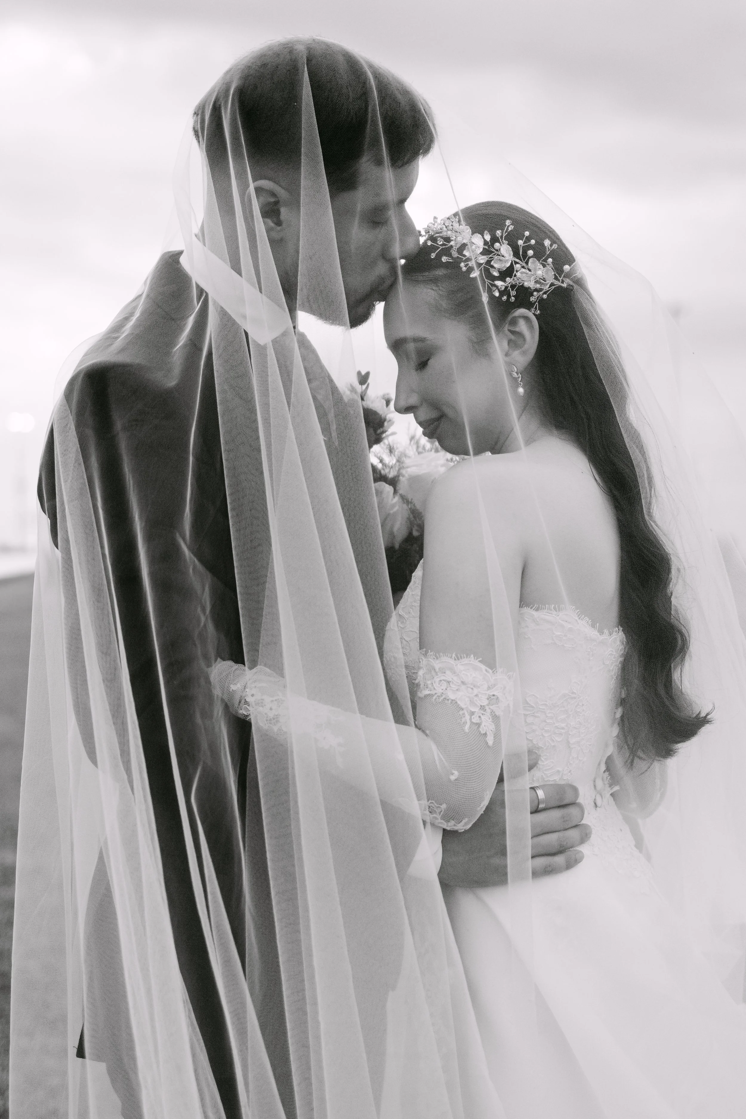 Una pareja de novios en un abrazo, la novia lleva un vestido de encaje y una diadema con flores, ambos con una expresión de felicidad, cubiertos por un velo de novios.