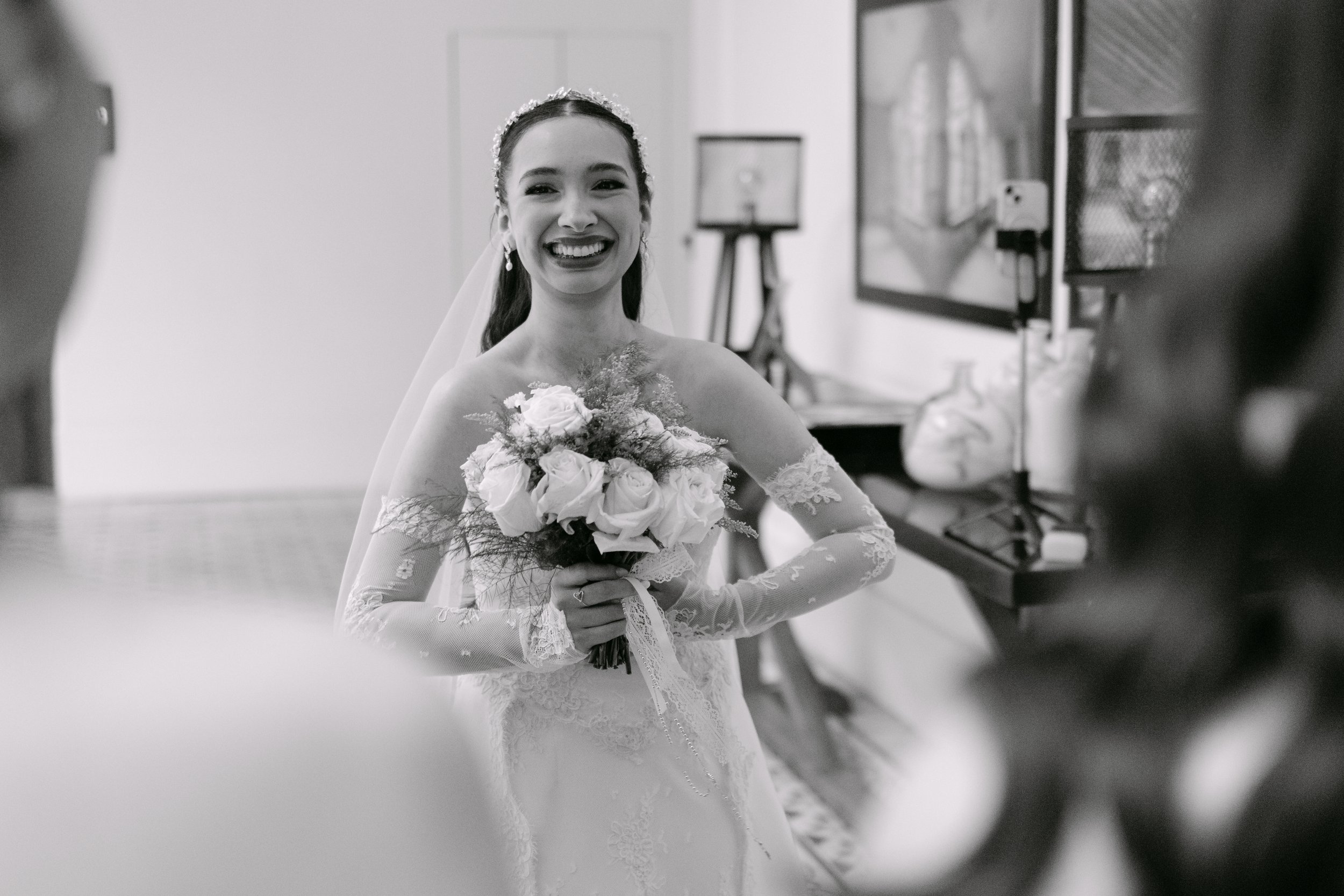 Mujer con vestido de novia y ramo de flores sonriente en una sala de eventos, vista en blanco y negro.