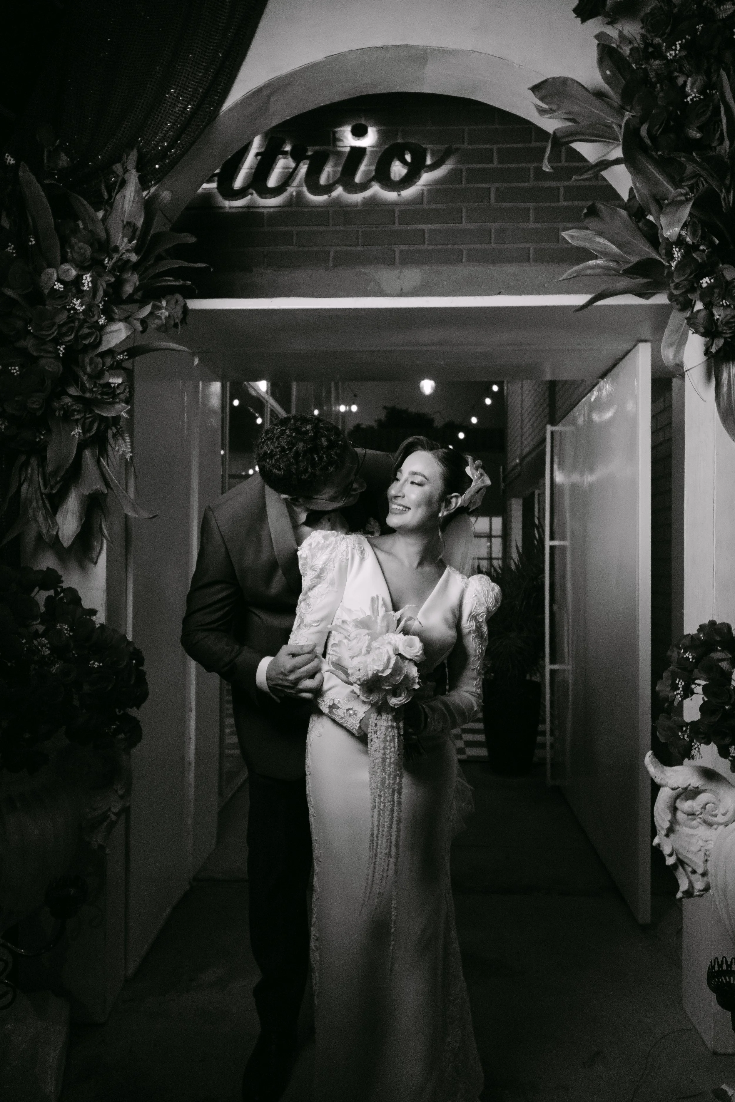 Una pareja de recién casados en la entrada de un lugar de celebración, ella con vestido de novia y él con traje, se miran y sonríen mientras se abrazan, decoraciones florales a los lados y señal luminosa que dice 'itrió' en el fondo.