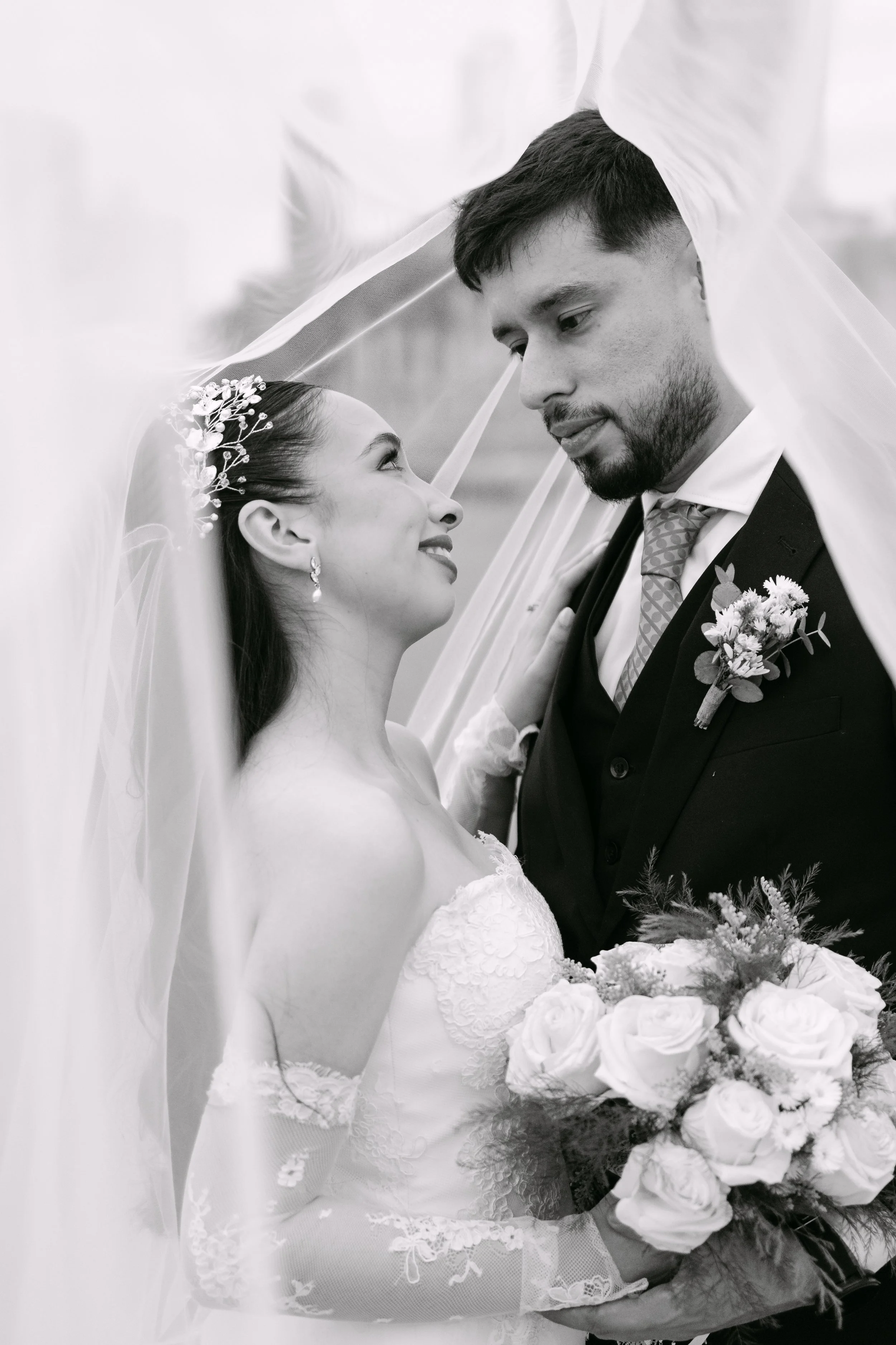 Pareja de recién casados bajo un velo de novia, ella con vestido de novia y ramo de flores blancas, él con traje formal y corbata, en una boda.