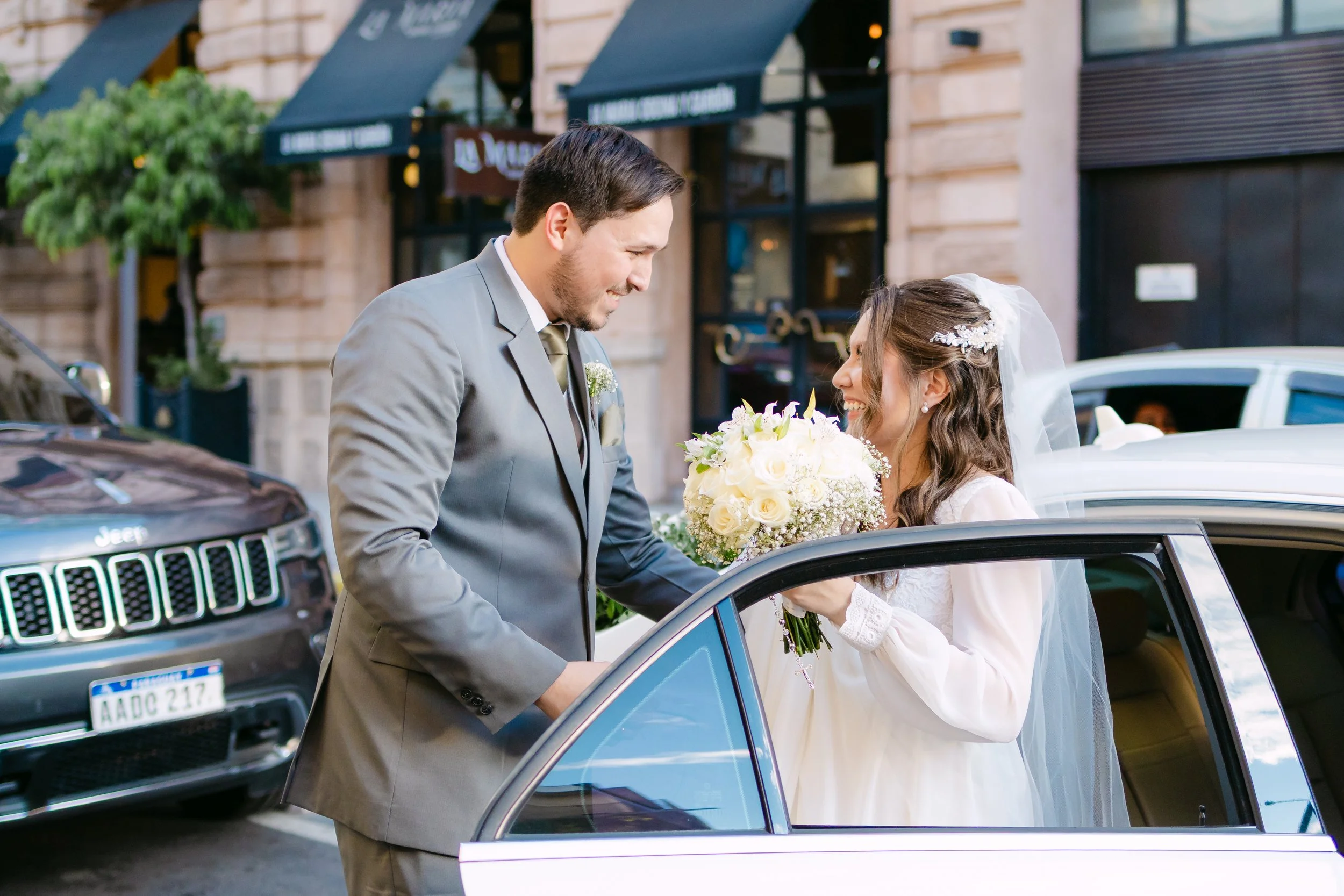 Novio y novia sonriendo y compartiendo un momento especial frente a un coche blanco en la calle, con la novia sosteniendo un ramo de flores blancas durante su boda.
