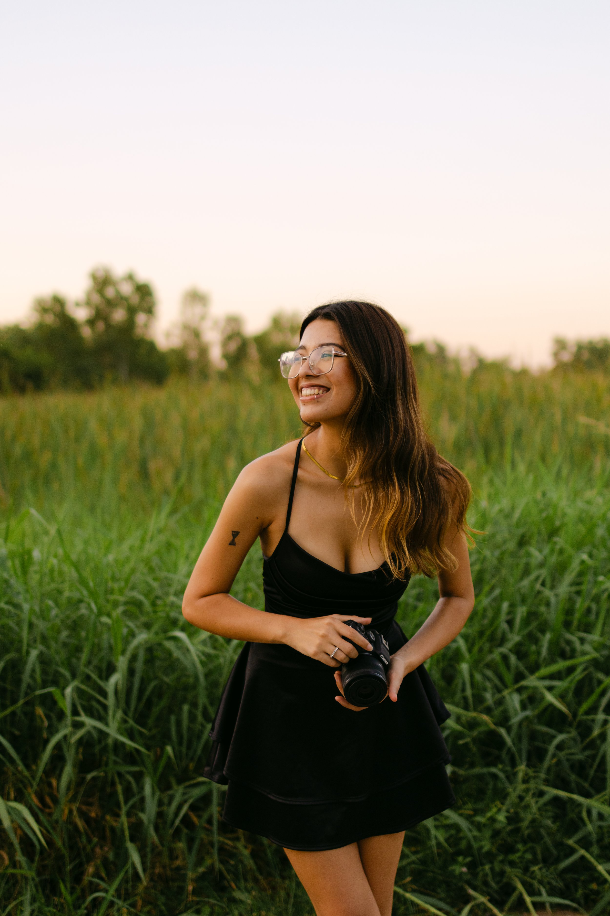 Joven mujer con gafas, vestido negro y tatuaje en el brazo, sonriendo y sosteniendo una cámara en un campo de hierba baja en la luz del atardecer.