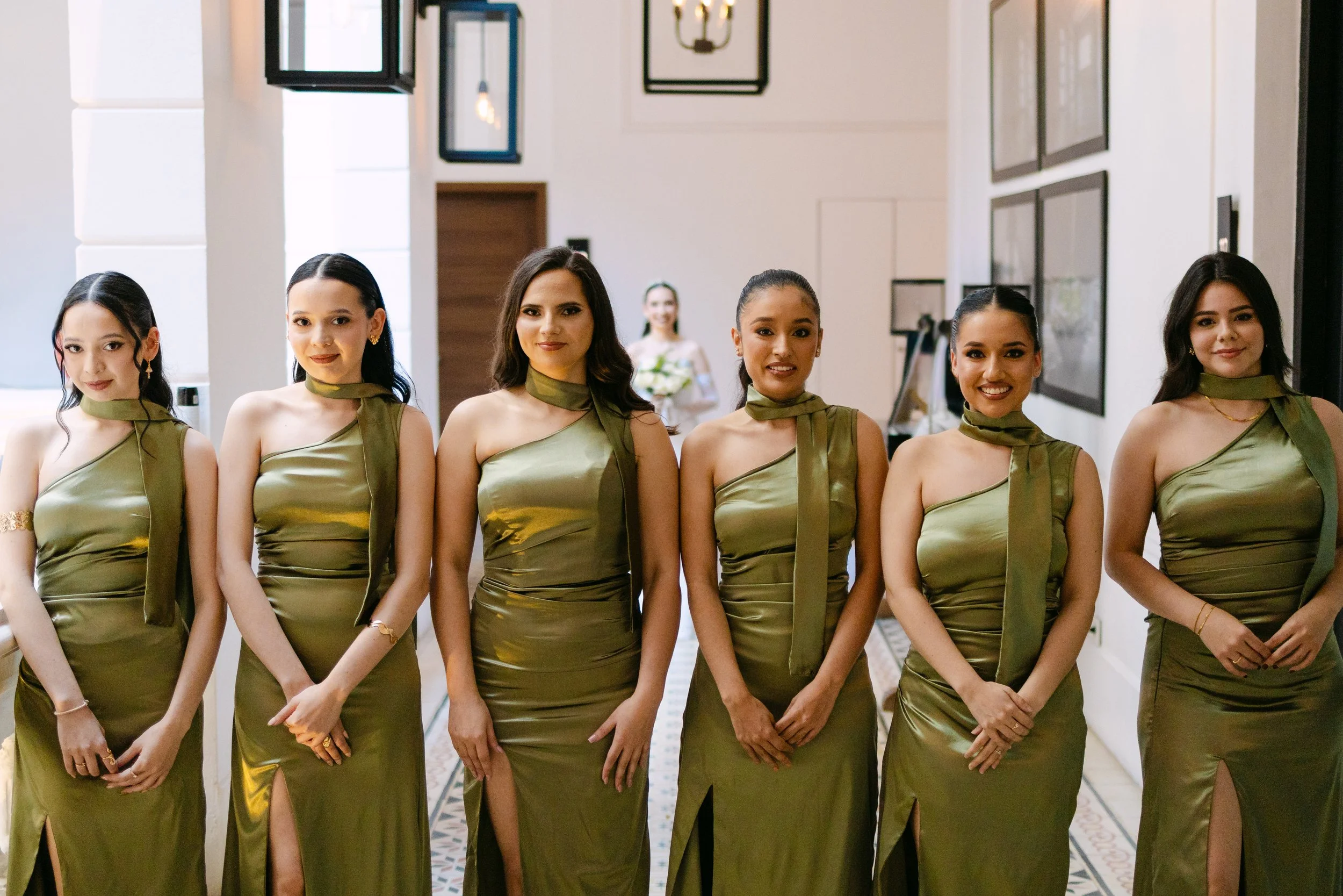 Grupo de seis mujeres vestidas con vestidos largos de color dorado en un evento, frente a una pared con cuadros y una mujer en el fondo con flores en la mano.