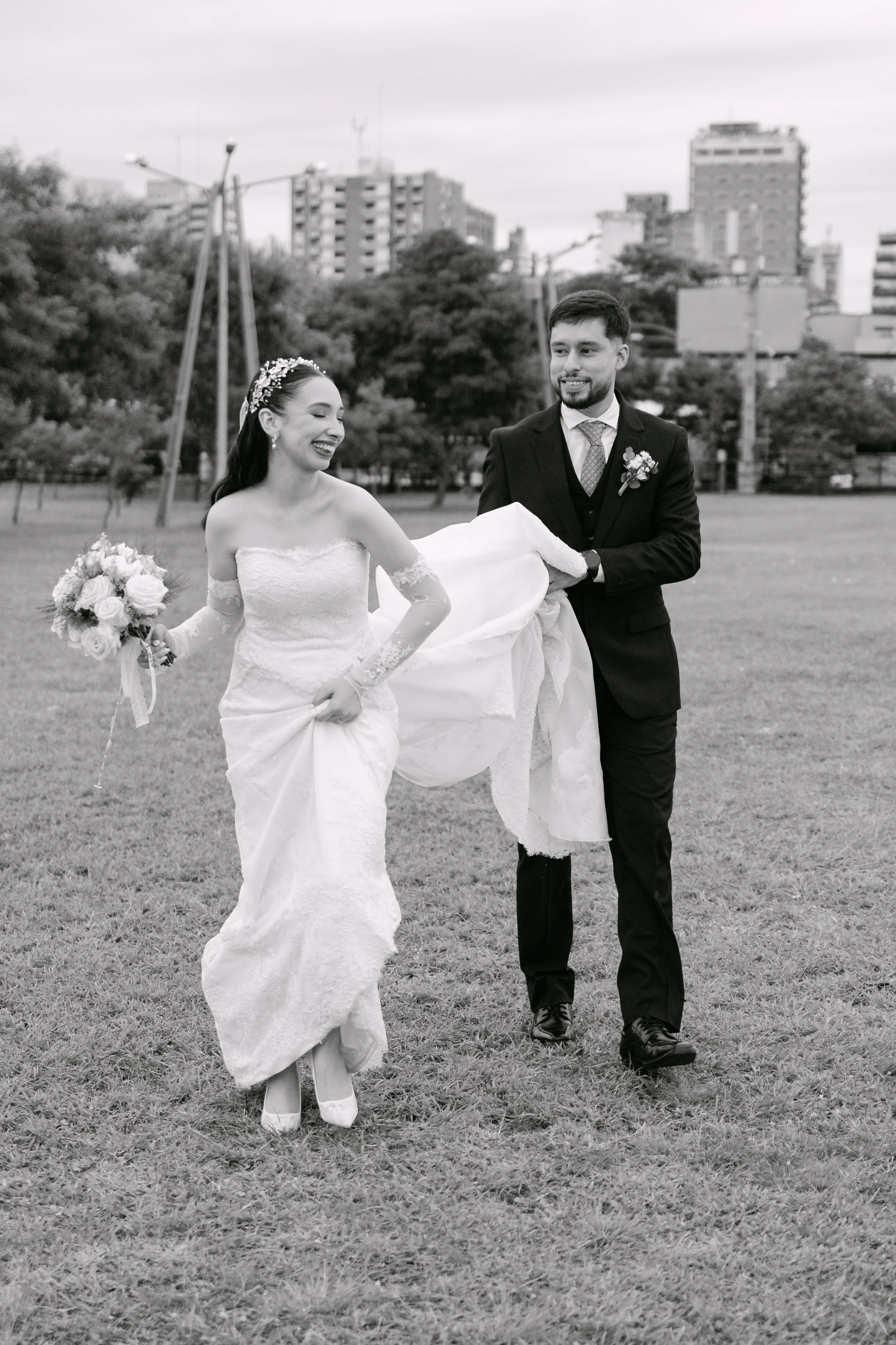 Pareja de recién casados, la mujer en vestido de novia con bouquet y el hombre en esmoquin, en un parque con edificios en el fondo, en blanco y negro.
