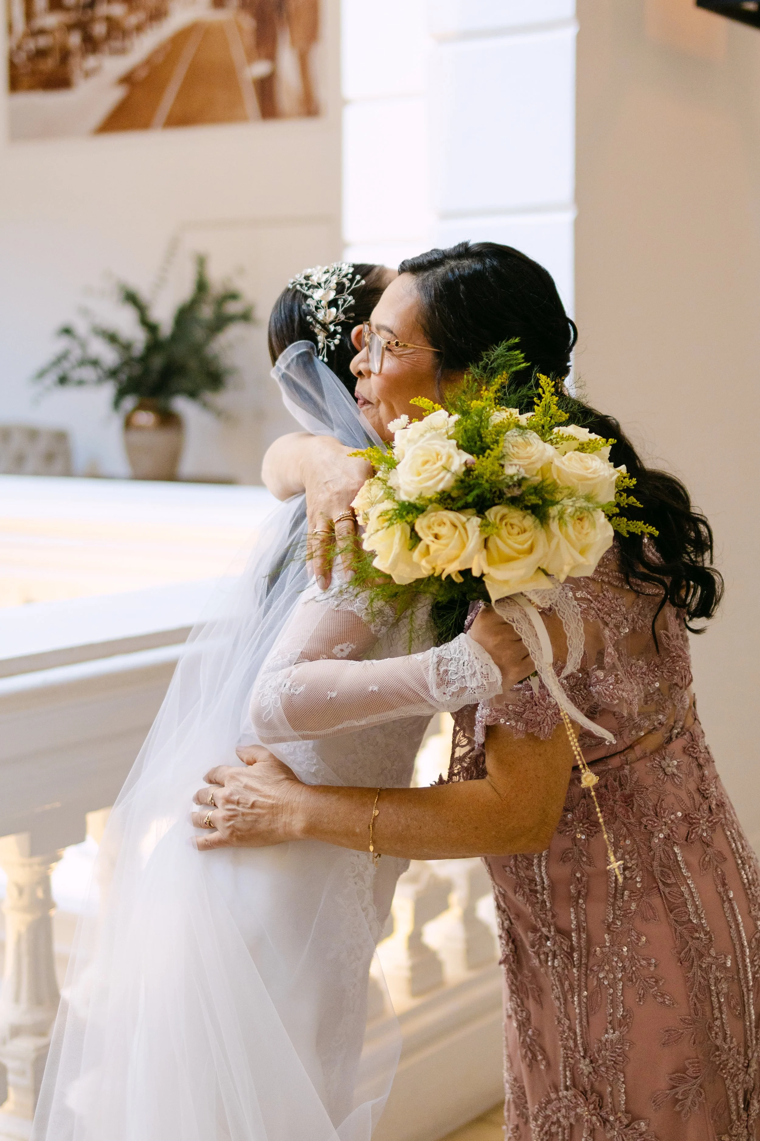 Una novia joven y una mujer adulta se abrazan, la novia sostiene un ramo de rosas blancas y verdes, ambas muestran cariño en un ambiente de interior con decoraciones elegantes.