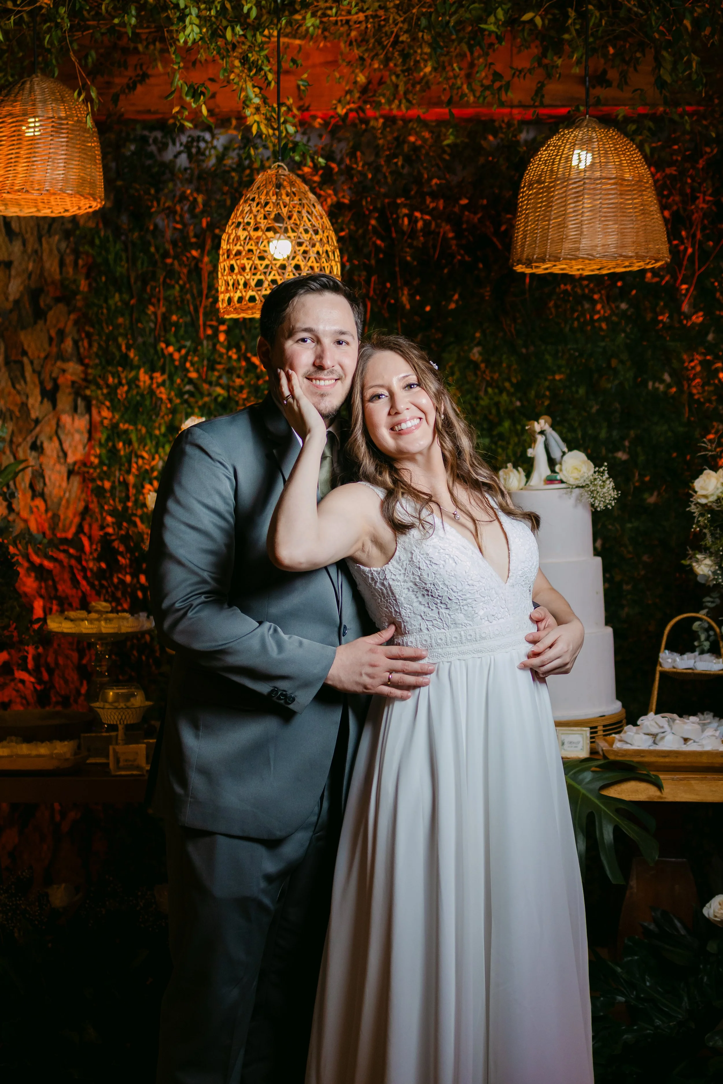 Pareja en una boda, el hombre lleva traje gris y la mujer vestido blanco de encaje, sonriendo y abrazados. Fondo con lámparas de mimbre, plantas y una torta de bodas decorada con flores.