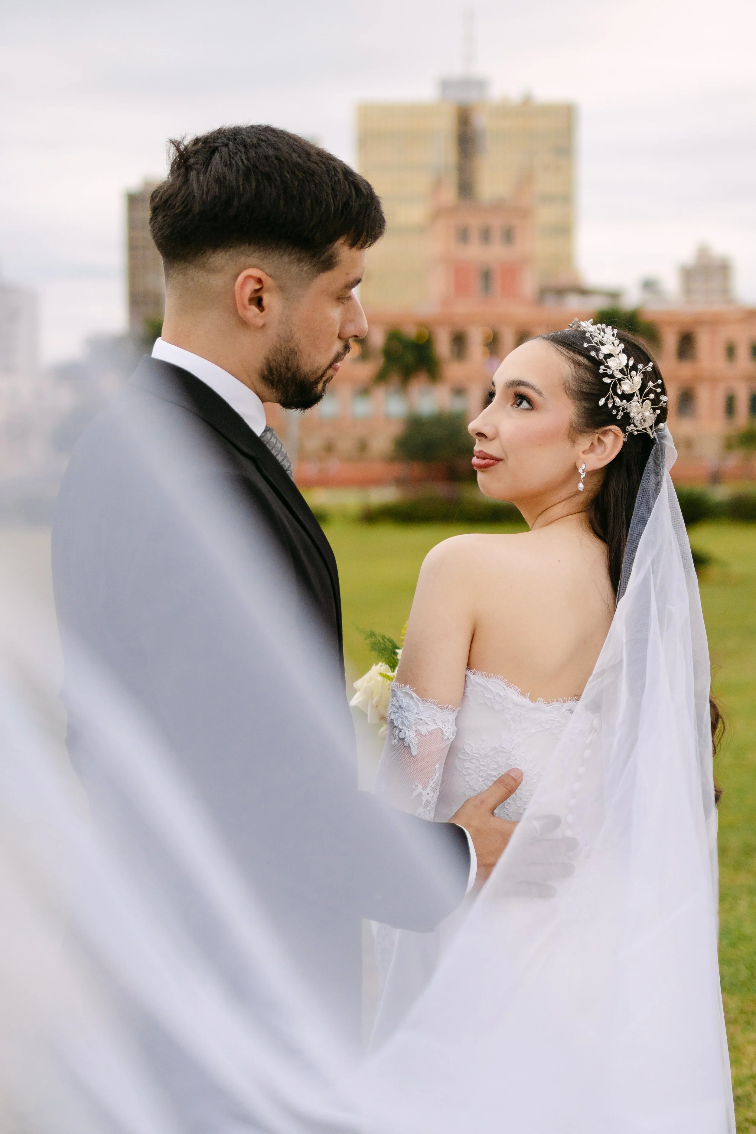 Pareja de novios en una ceremonia de bodas al aire libre, con edificios en el fondo, la novia lleva un vestido de encaje y un adorno en la cabeza, y el novio viste un saco y corbata.