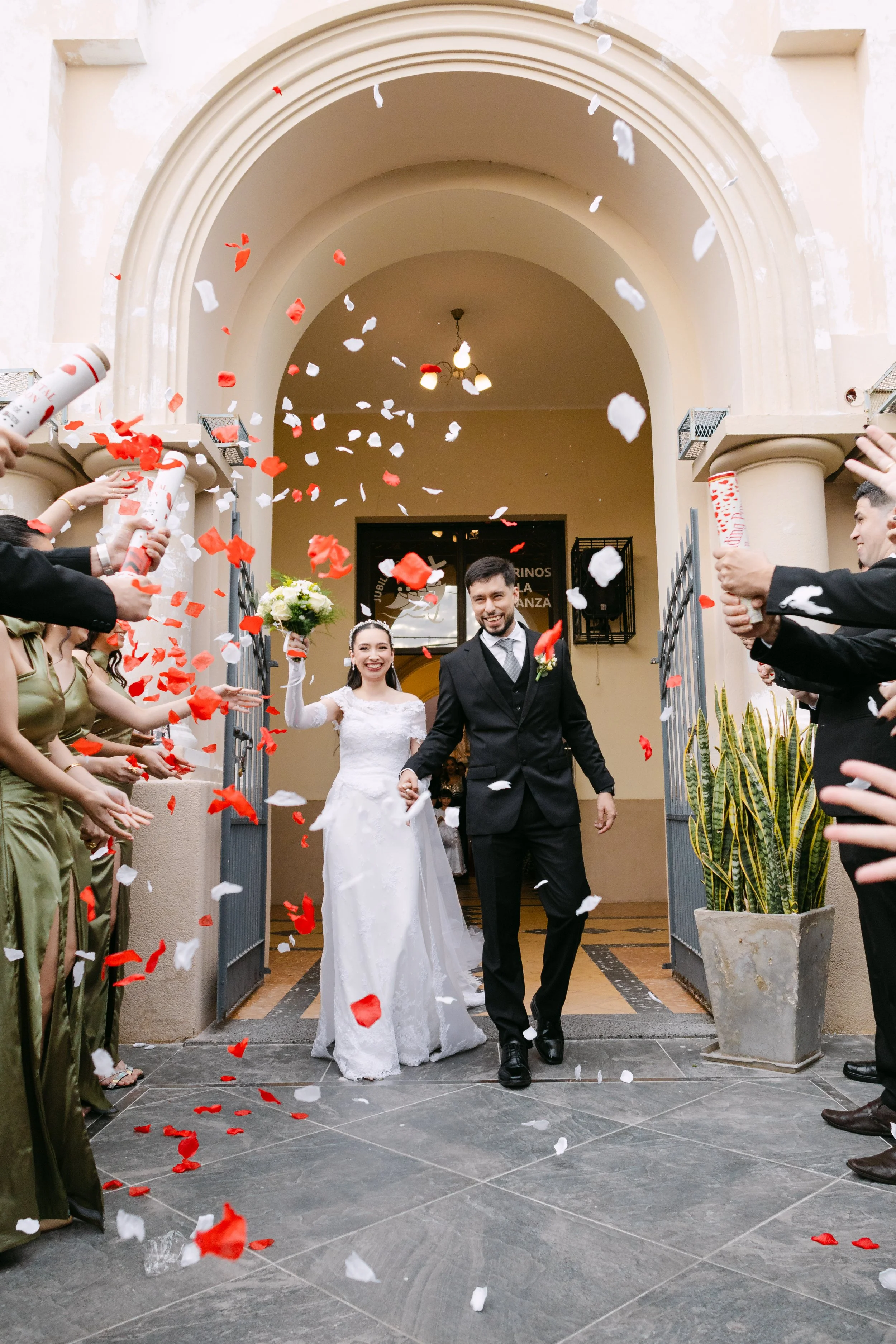 Pareja de recién casados saliendo entre confeti rojo y blanco en la entrada de un edificio, rodeados de amigos y familiares, en medio de una celebración.