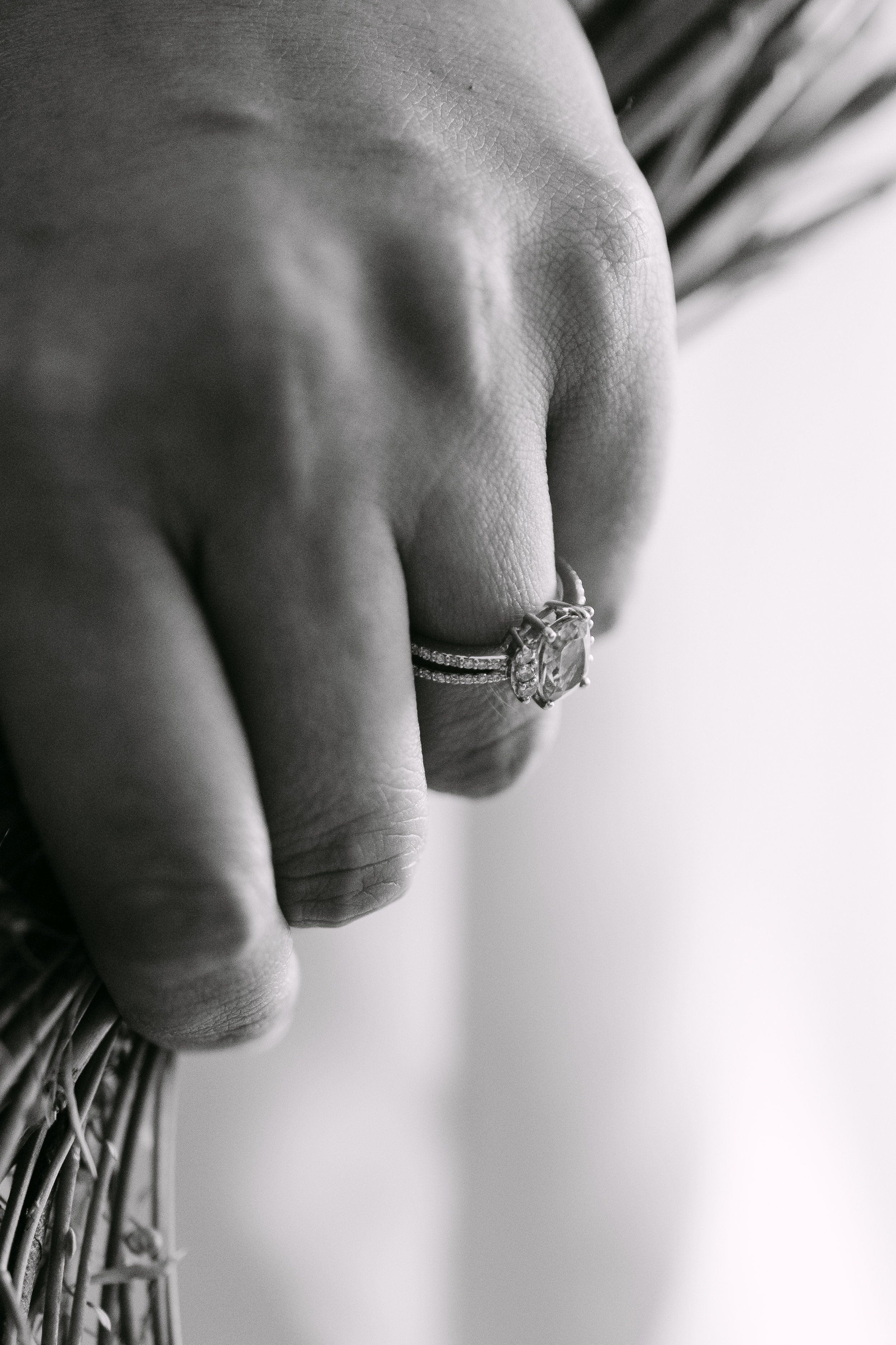 Primer plano de una mano con un anillo en el dedo medio, la mano descansa sobre un cesto de mimbre o ratán en tonos de gris, en un fondo difuso.