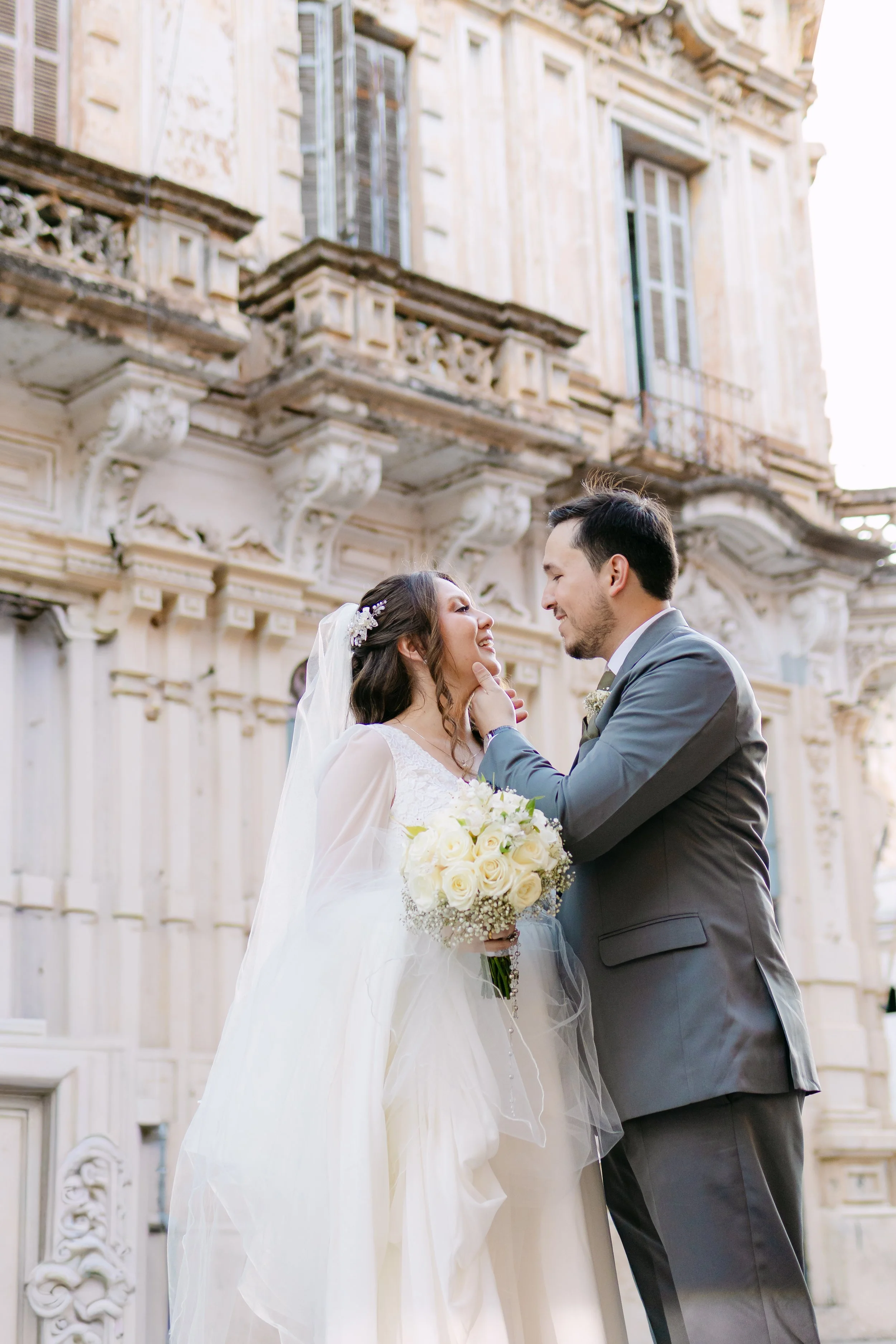 Pareja de recién casados sonrientes, la novia con vestido blanco y ramo de rosas blancas, el novio con traje gris, frente a un edificio antiguo.
