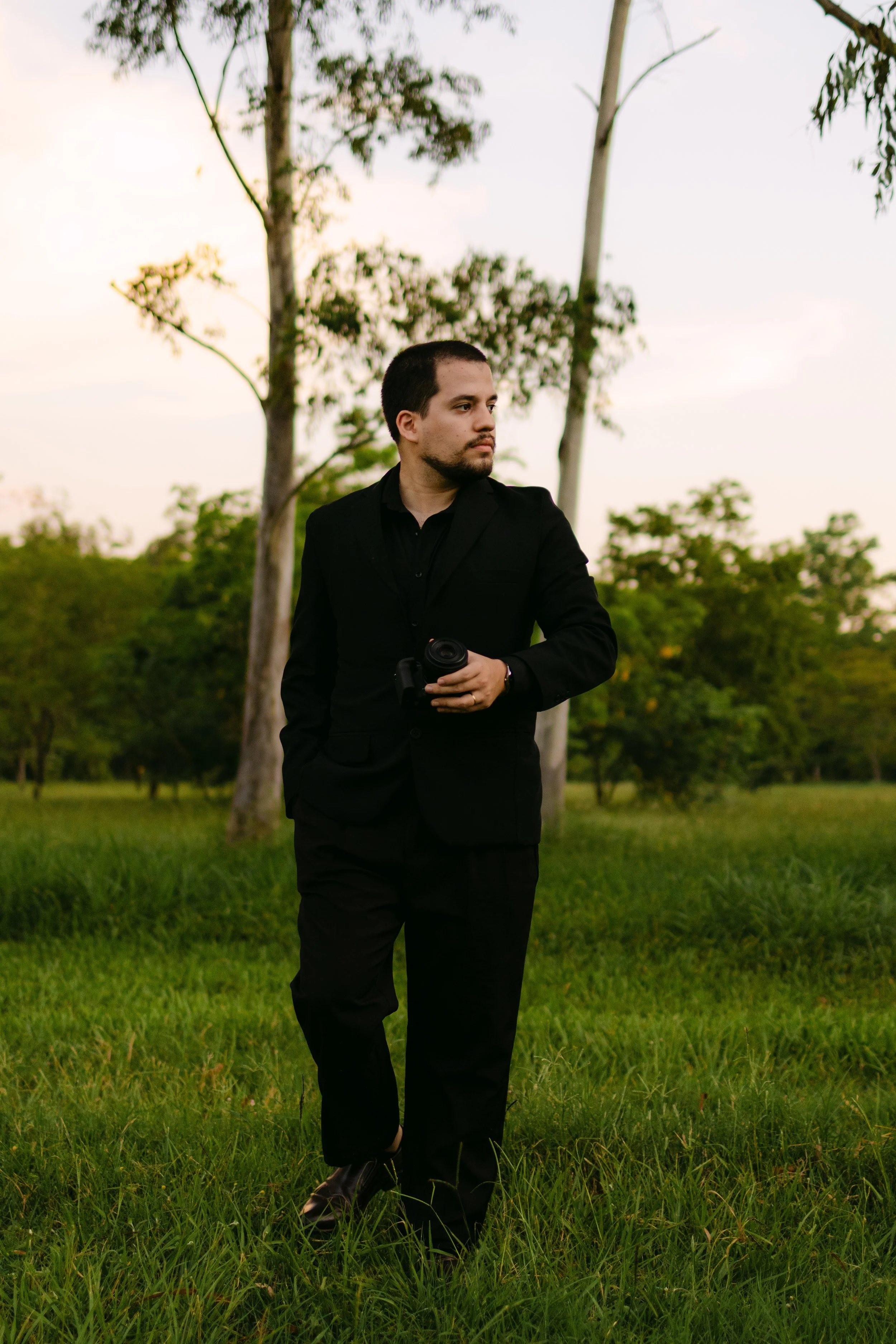 Un hombre vestido con traje negro caminando por un campo con árboles, sosteniendo una cámara en la mano, en un entorno natural y al atardecer.