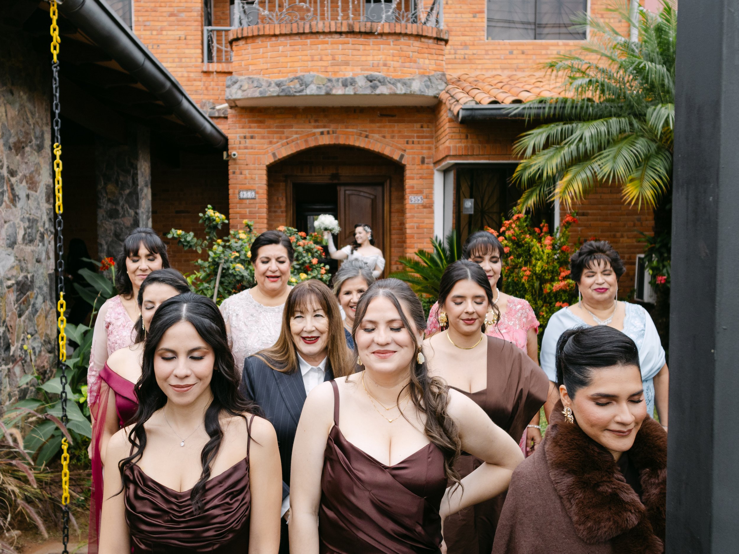 Grupo de mujeres en un evento social, con casas de ladrillo y plantas en el fondo.