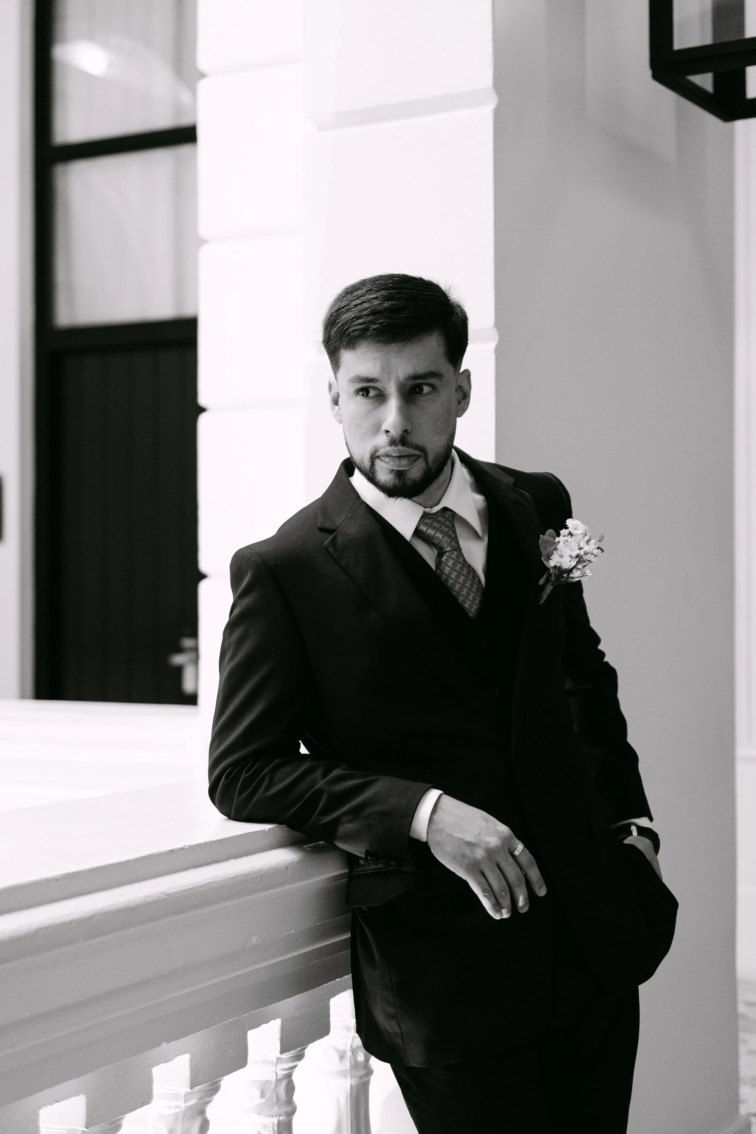 Hombre con barba y peinado de cabello oscuro, vestido con saco oscuro, camisa blanca, corbata y flor en el saco, posando junto a una barandilla en un entorno interior moderno y bien iluminado.